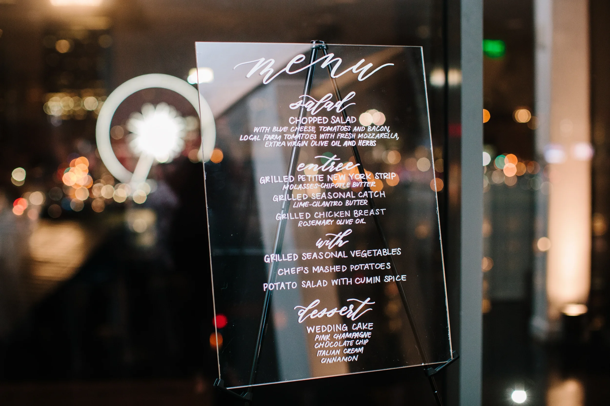 Bring a ultra-modern touch to your tabletop design with a clear acrylic wedding menu sign. Featuring crisp white hand-lettering, this sleek transparent menu stands out beautifully against twinkling city night lights, making it perfect for a luxury ur