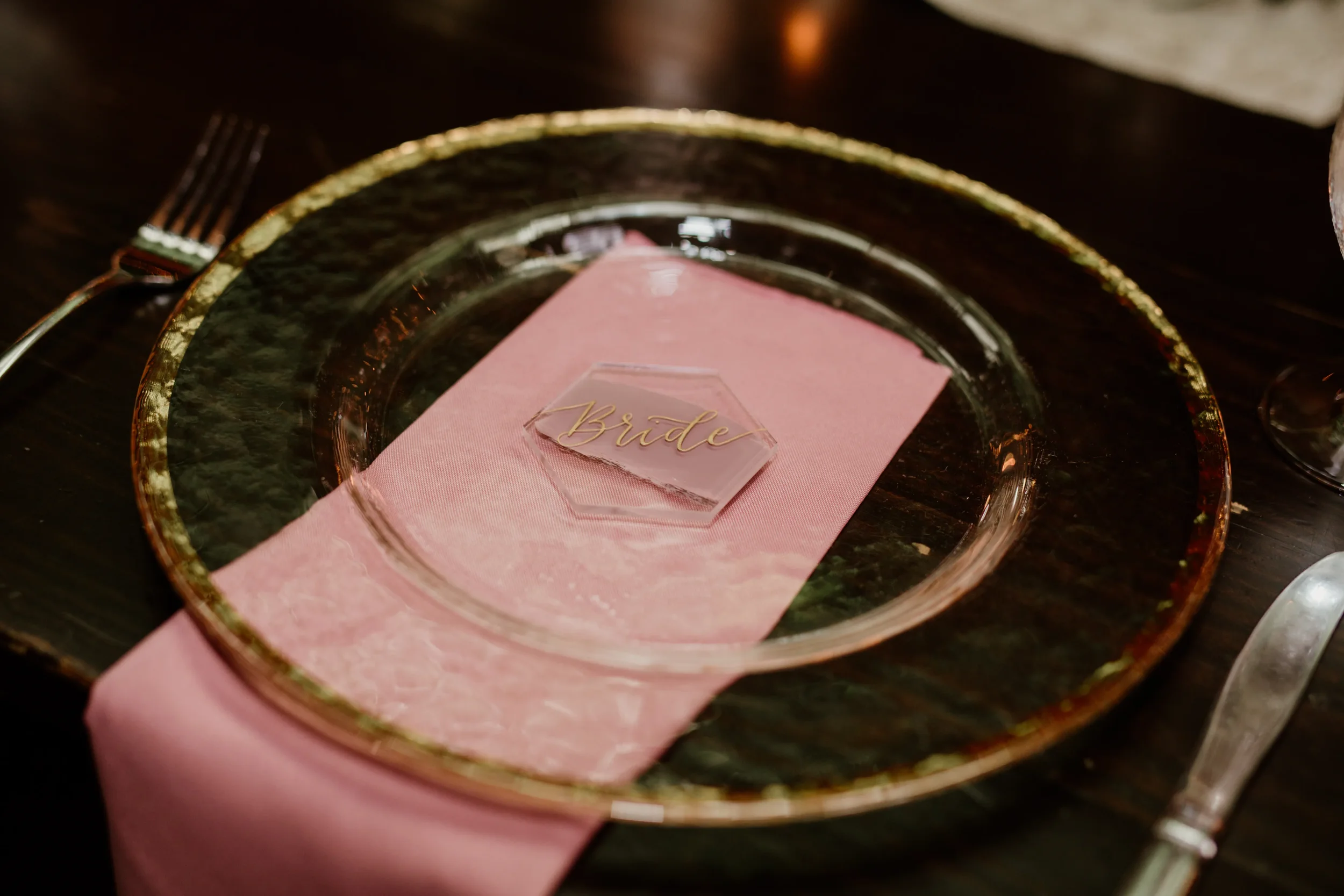 Elevate your fine-art tablescape with custom geometric clear acrylic place cards. Featuring fluid gold script calligraphy, this hexagon name card is layered over a dusty pink napkin and a textured glass charger plate to create a deeply romantic, high