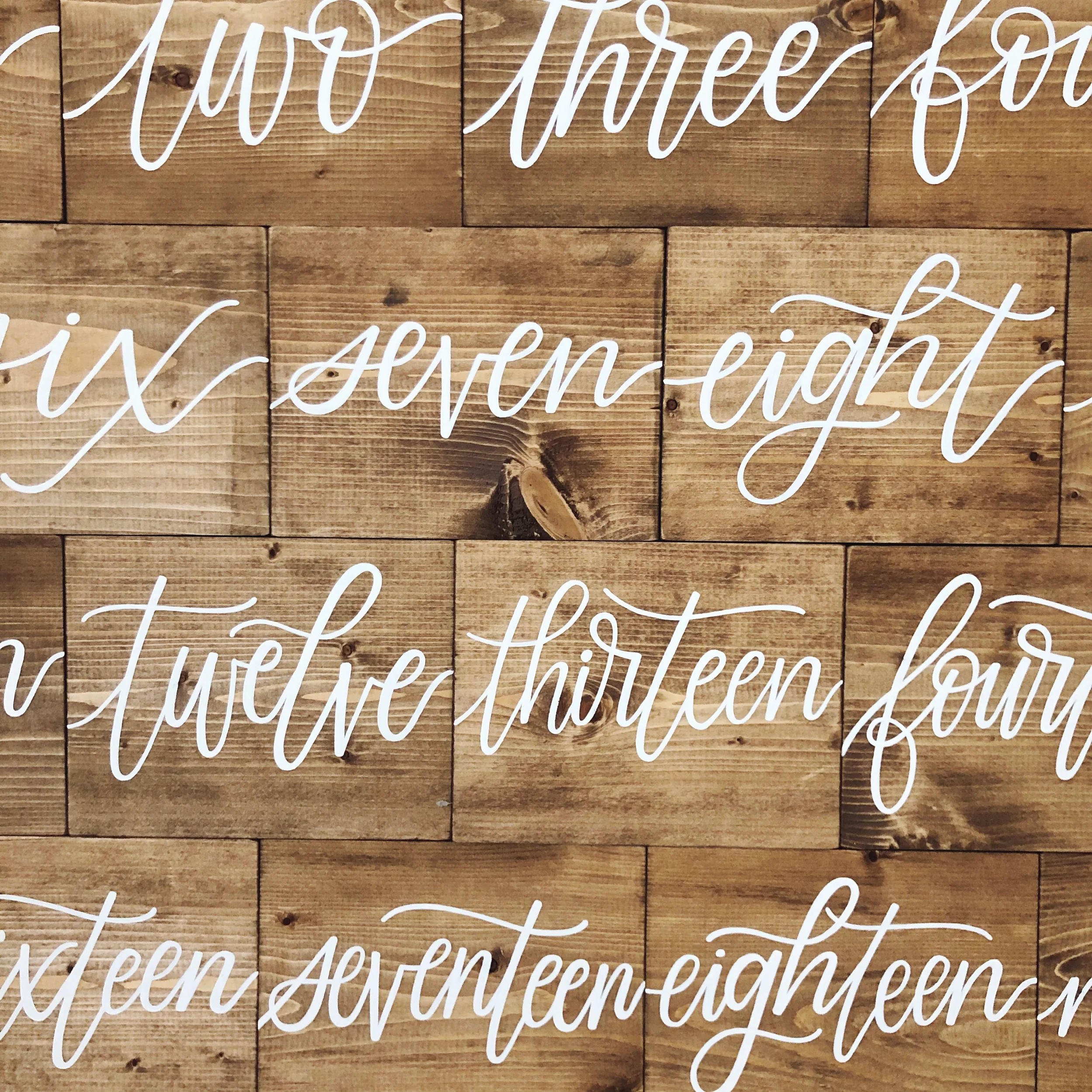 Add warm rustic charm to your reception with a freestanding stained wood block table number, lettered in bold white cursive script.