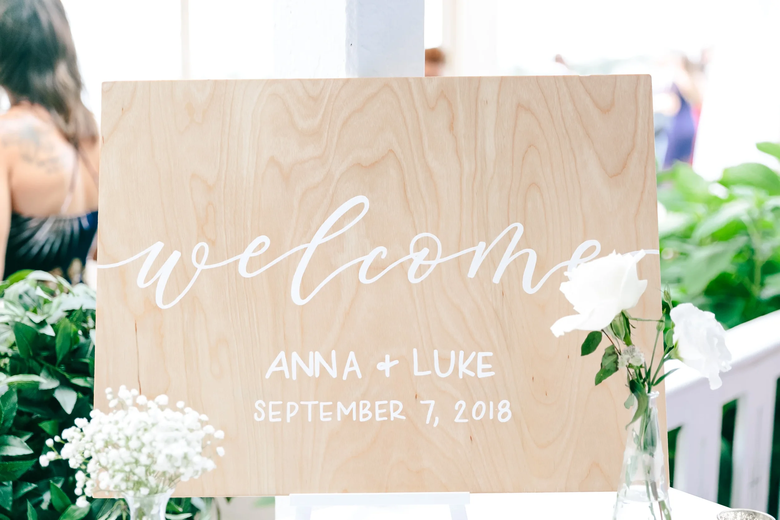 Horizontal tabletop natural wood wedding welcome sign hand-lettered in white ink with the couple's names and wedding date.