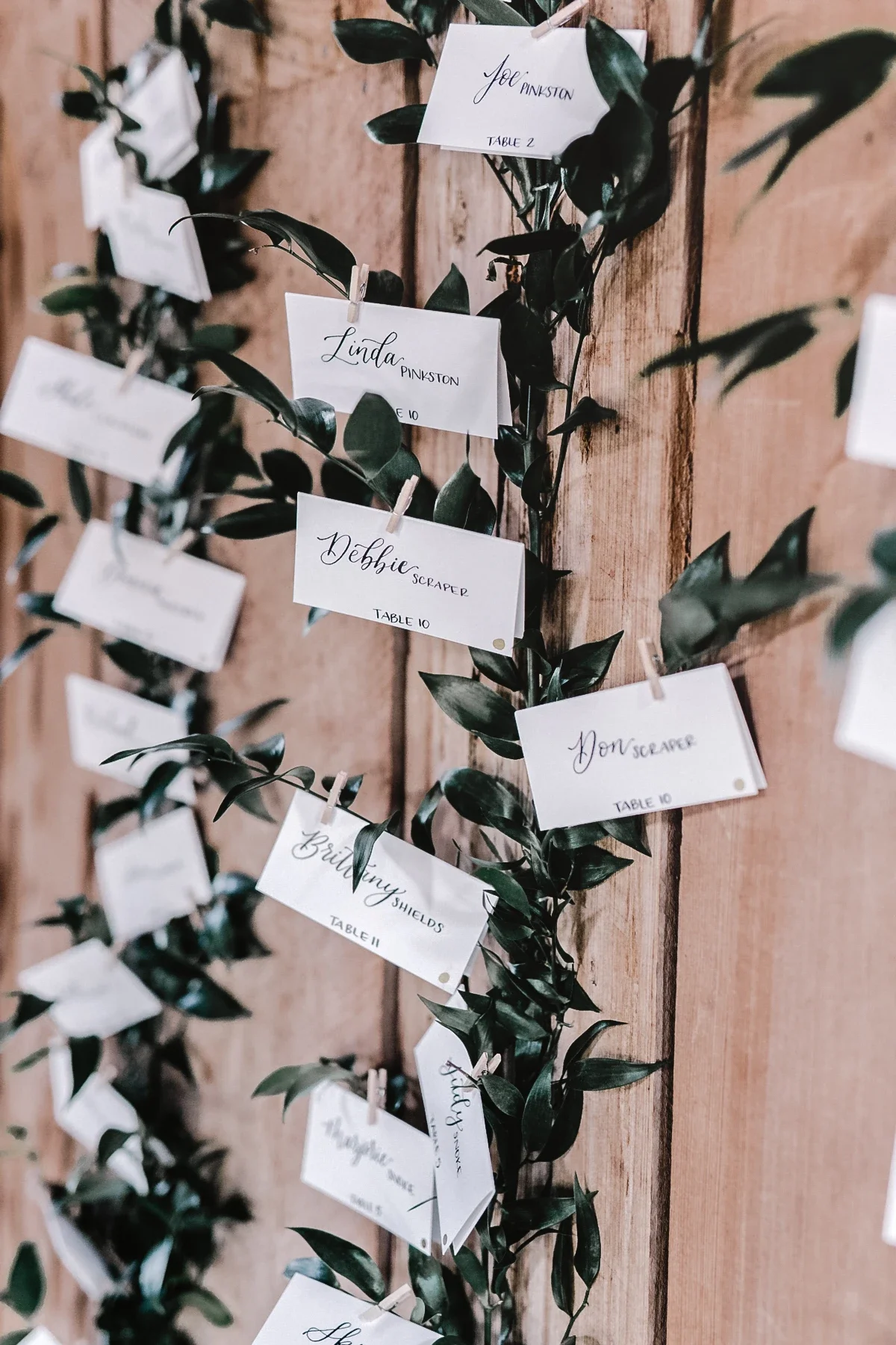Hand-lettered white escort cards clipped with mini clothespins onto greenery vines against a wood plank backdrop.