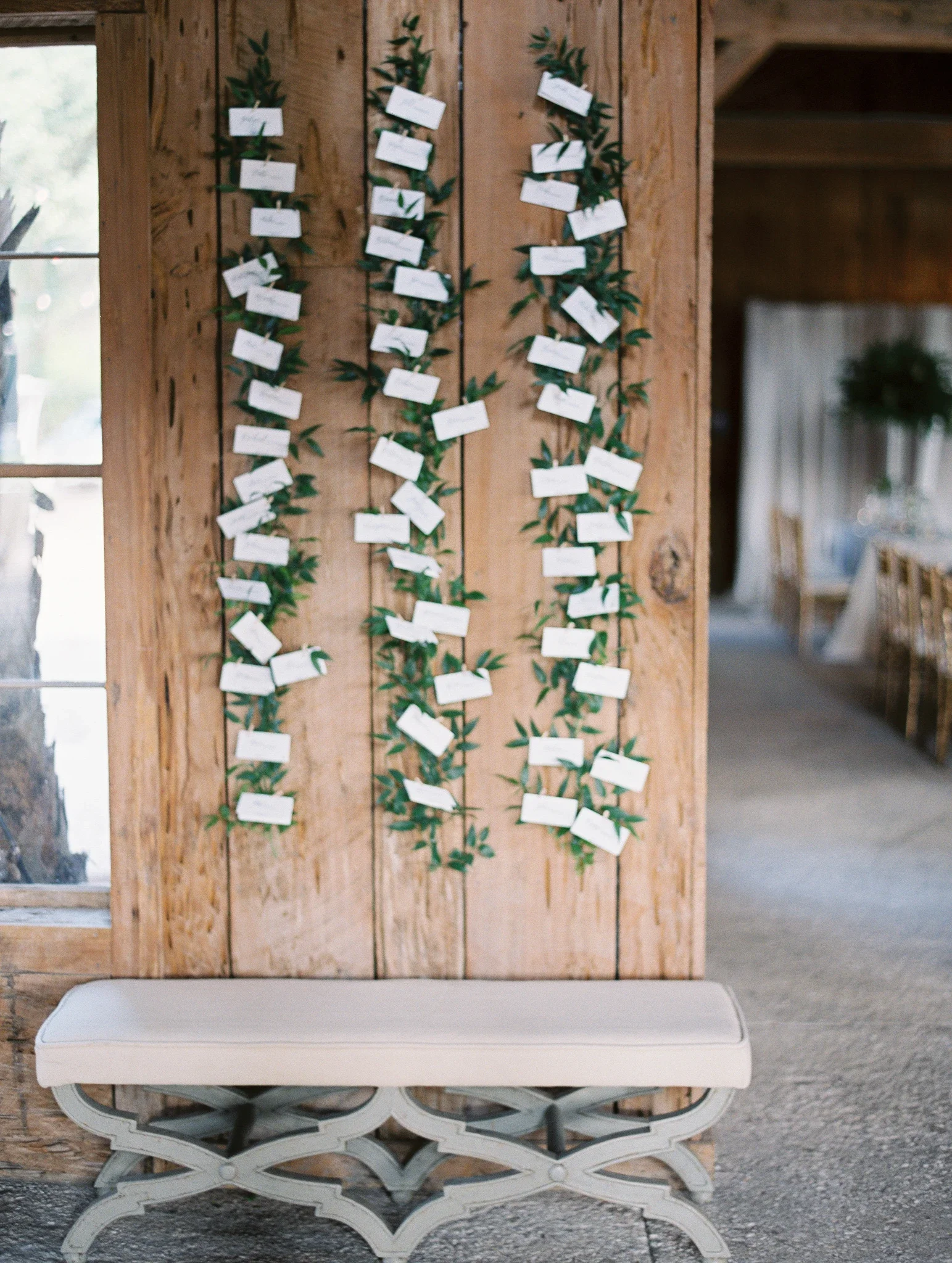 Create an organic, garden-inspired arrival experience with a greenery-draped escort card display. Featuring white hand-lettered cards attached to trailing vines with mini clothespins against rustic wood paneling, this seating wall adds natural elegan