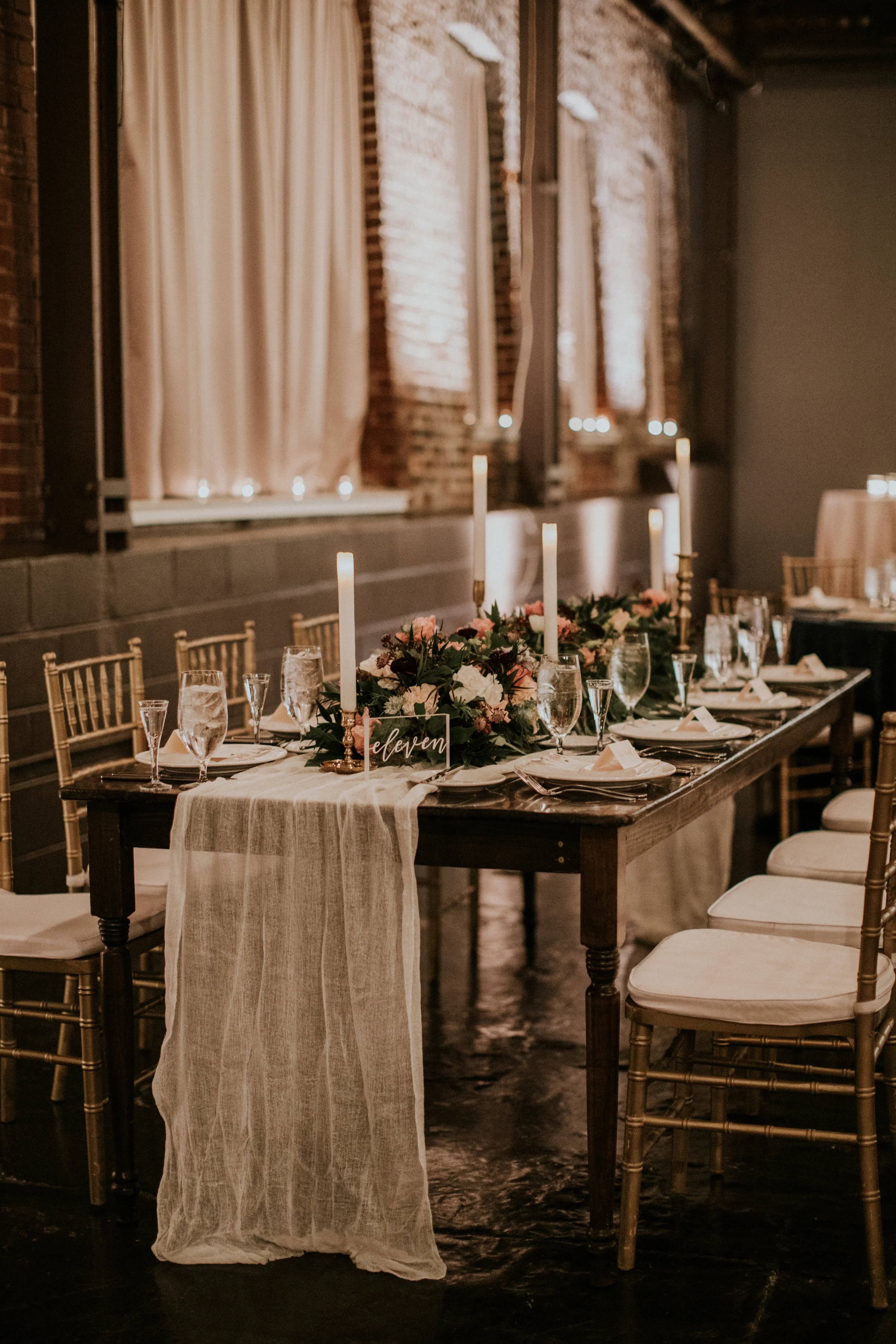 Create a romantic atmosphere with custom acrylic wedding table numbers featuring elegant white script calligraphy. This clear lucite table sign reading "eleven" is beautifully styled on a dark wood farm table with an organic white gauze runner, lush 