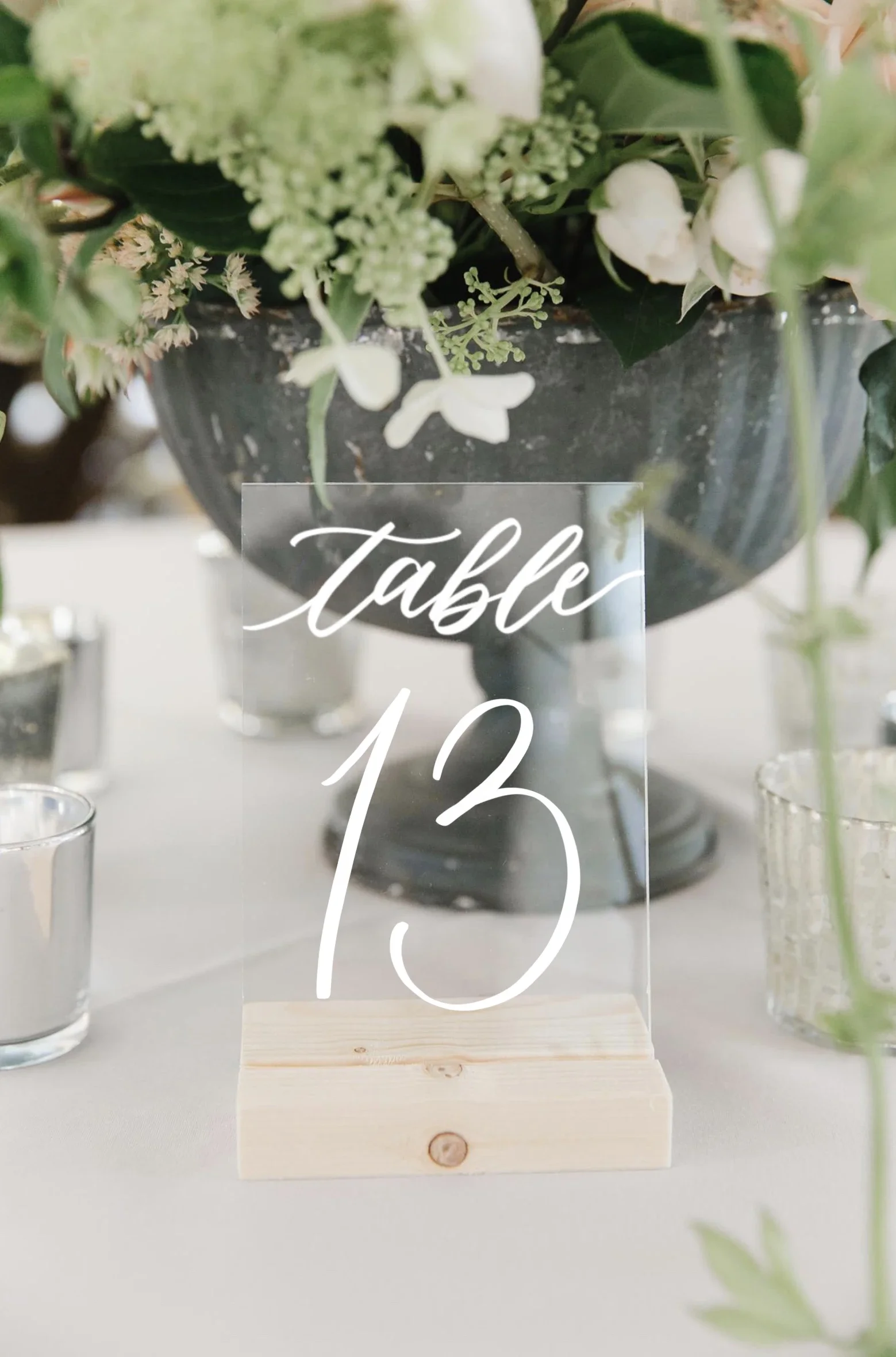 Perfectly suited for modern or organic tablescapes, this clear acrylic table number features crisp white calligraphy mounted on a natural wood block base. Its transparent design blends effortlessly into lush floral centerpieces.