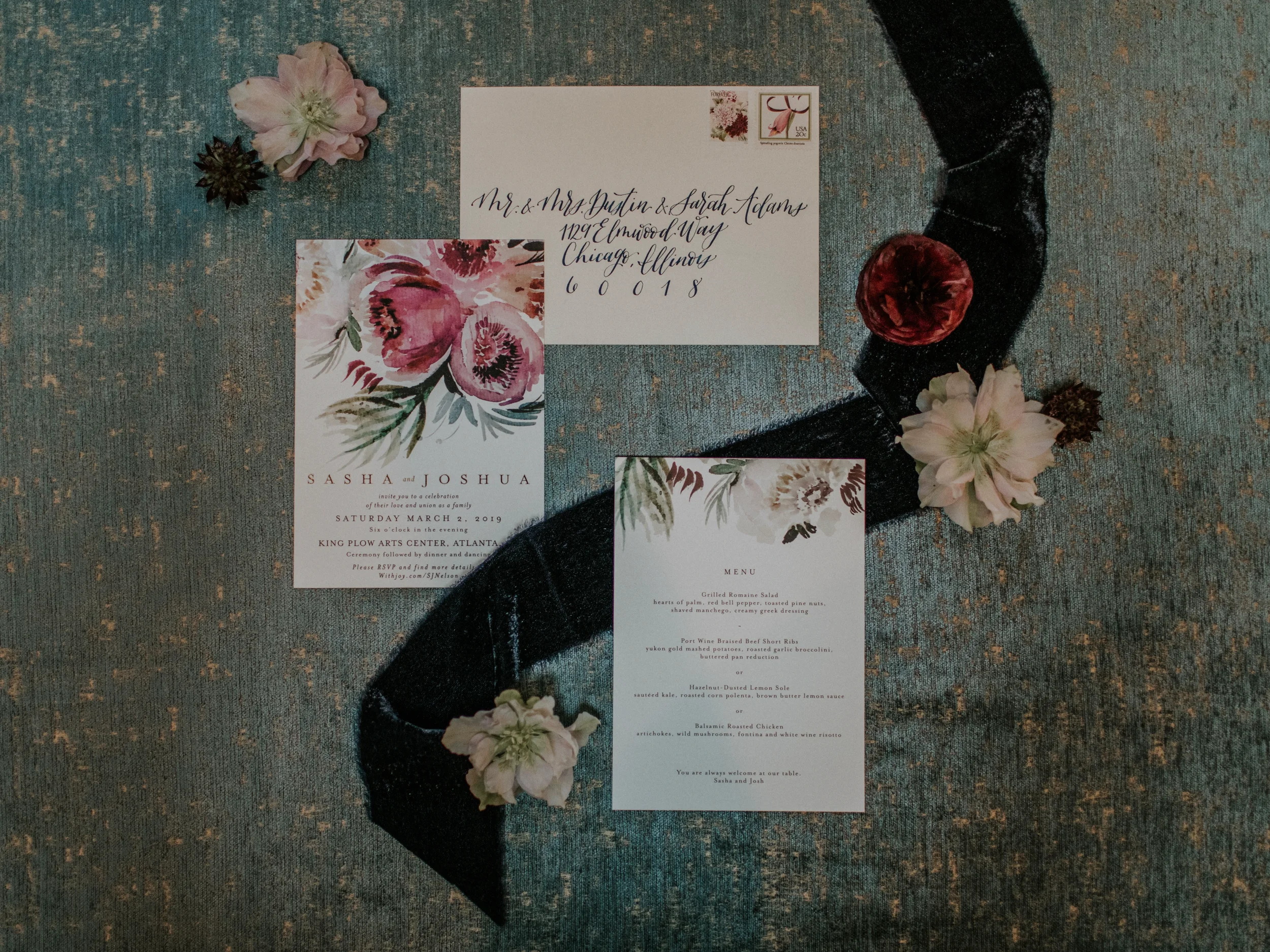Flat lay overview of a wedding stationery suite featuring a hand-addressed calligraphy envelope, a floral watercolor wedding invitation, and a menu card arranged on a textured teal backdrop with a navy velvet ribbon and loose flowers.