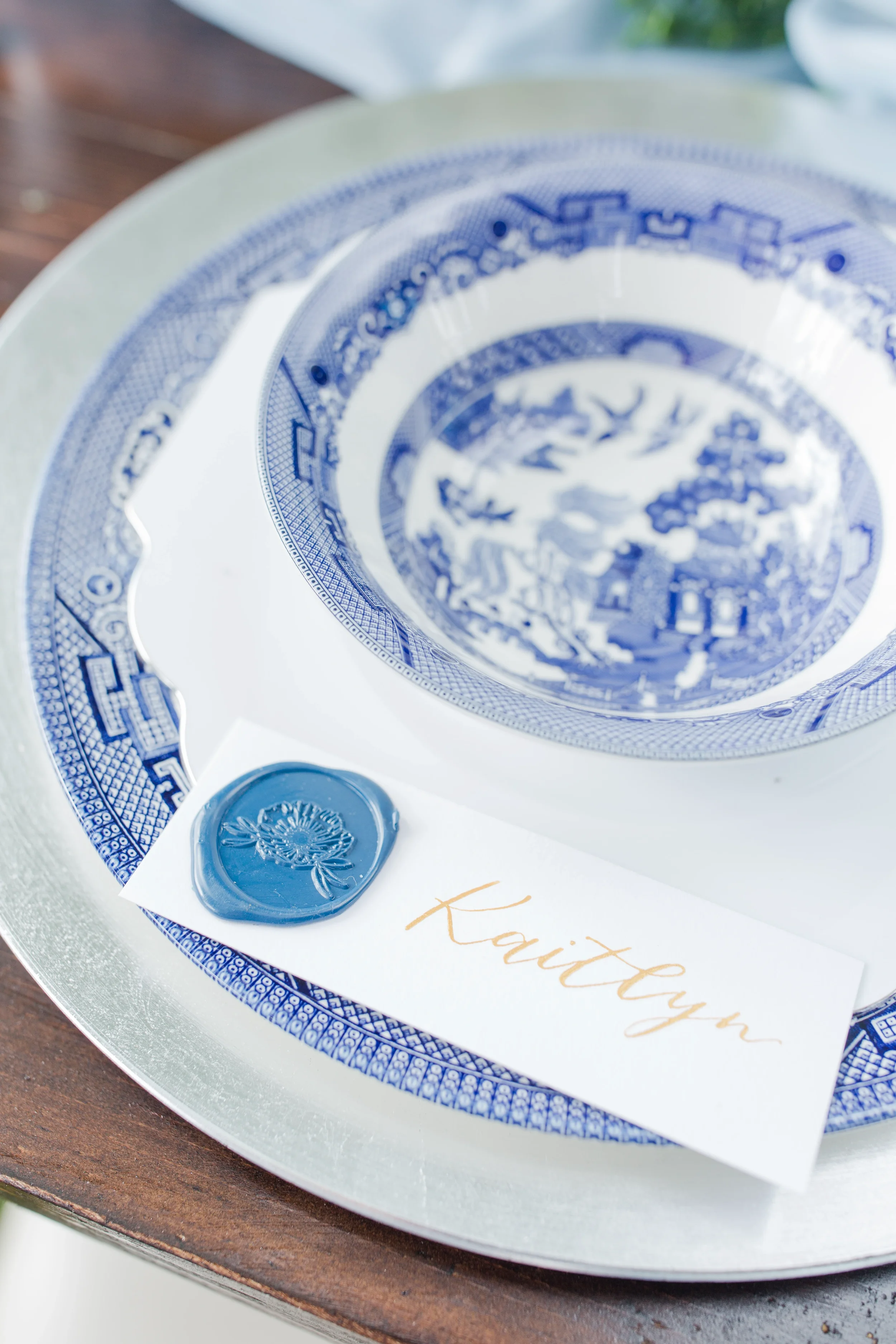 Layer classic pattern work into your reception dinner with a blue willow china place setting. Topped with a personalized place card featuring a custom blue wax seal and gold calligraphy script, this table design bridges traditional vintage charm with