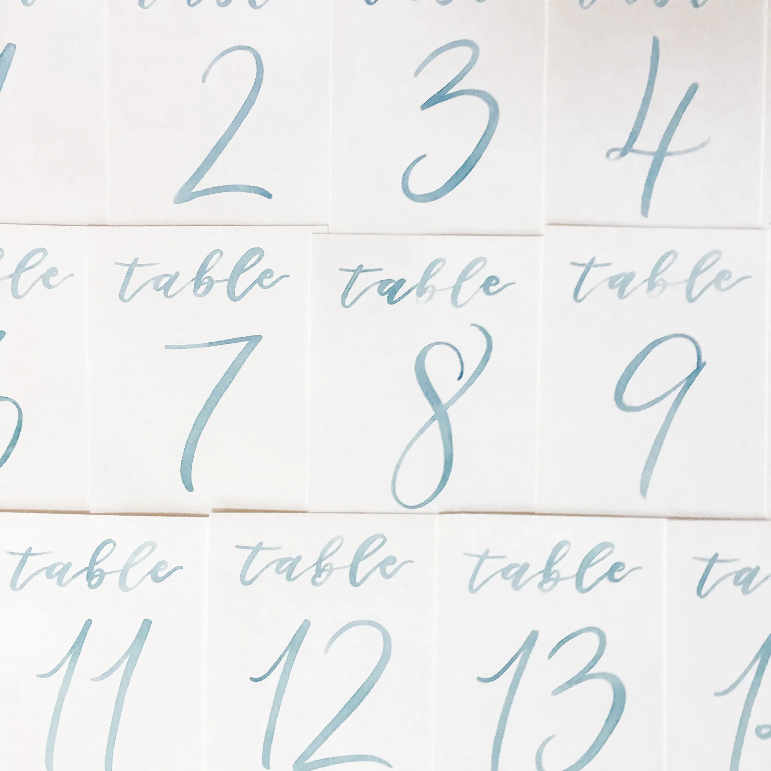 Light dusty blue handwritten script table numbers on clean white paper cards.