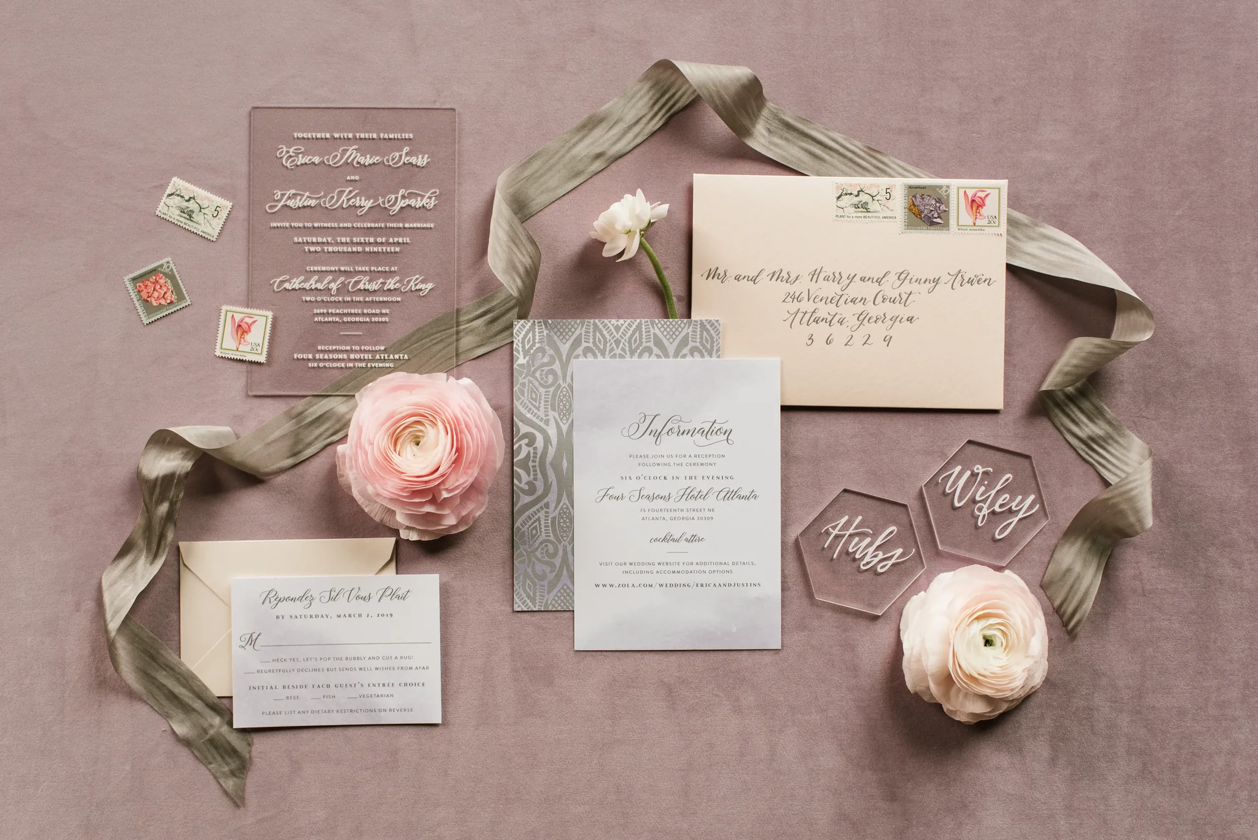 Clear acrylic wedding invitation suite with gold script, styled on mauve fabric with olive green silk ribbon and pink ranunculus flowers.