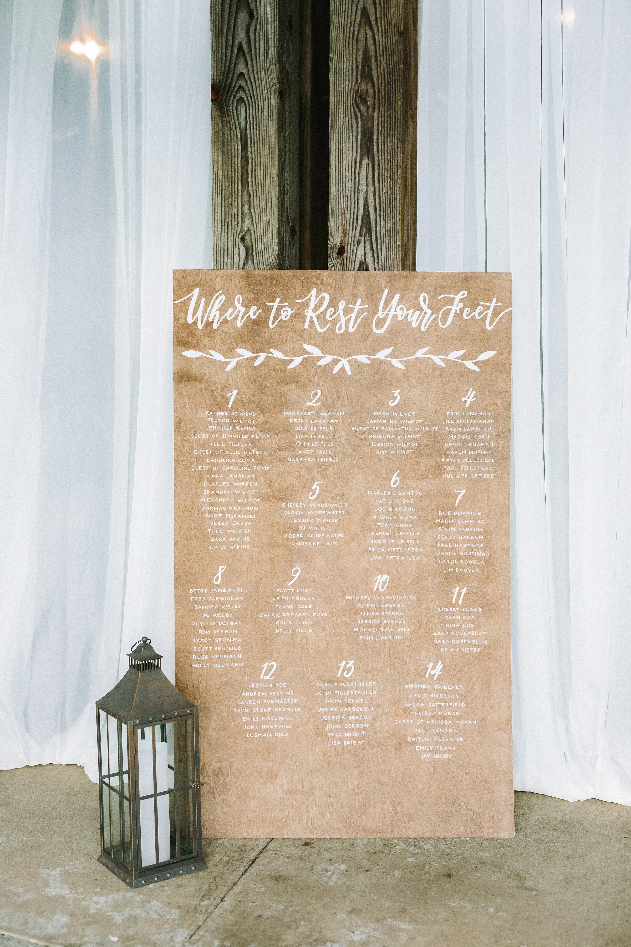 Guide your guests with warm, playful phrasing using a custom plywood seating chart. Hand-lettered in crisp white calligraphy under the header "Where to Rest Your Feet," this freestanding wood panel brings a relaxed yet refined aesthetic to barn or ga