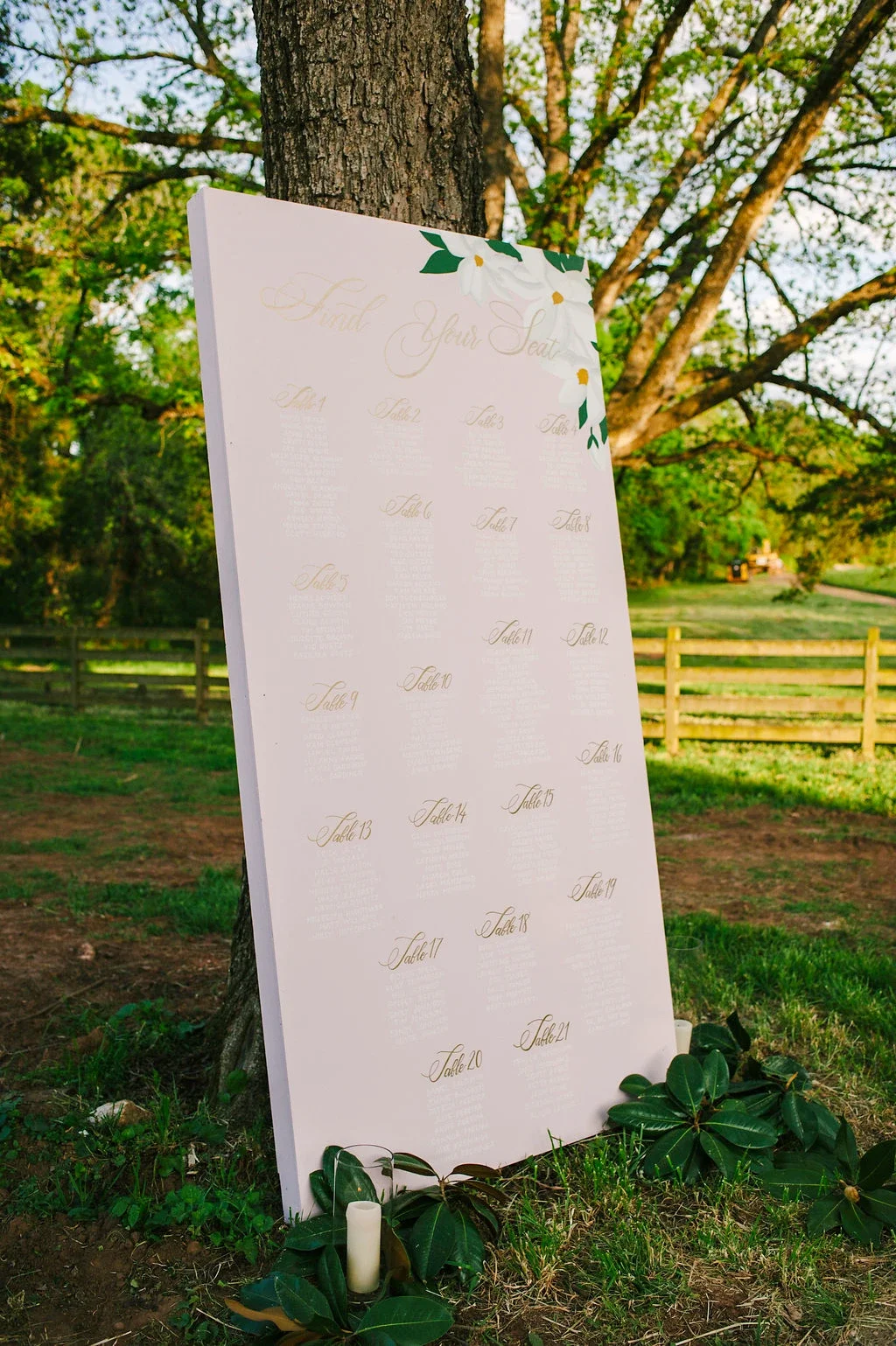 Standing blush pink wedding seating chart board placed outdoors on grass next to a large tree.