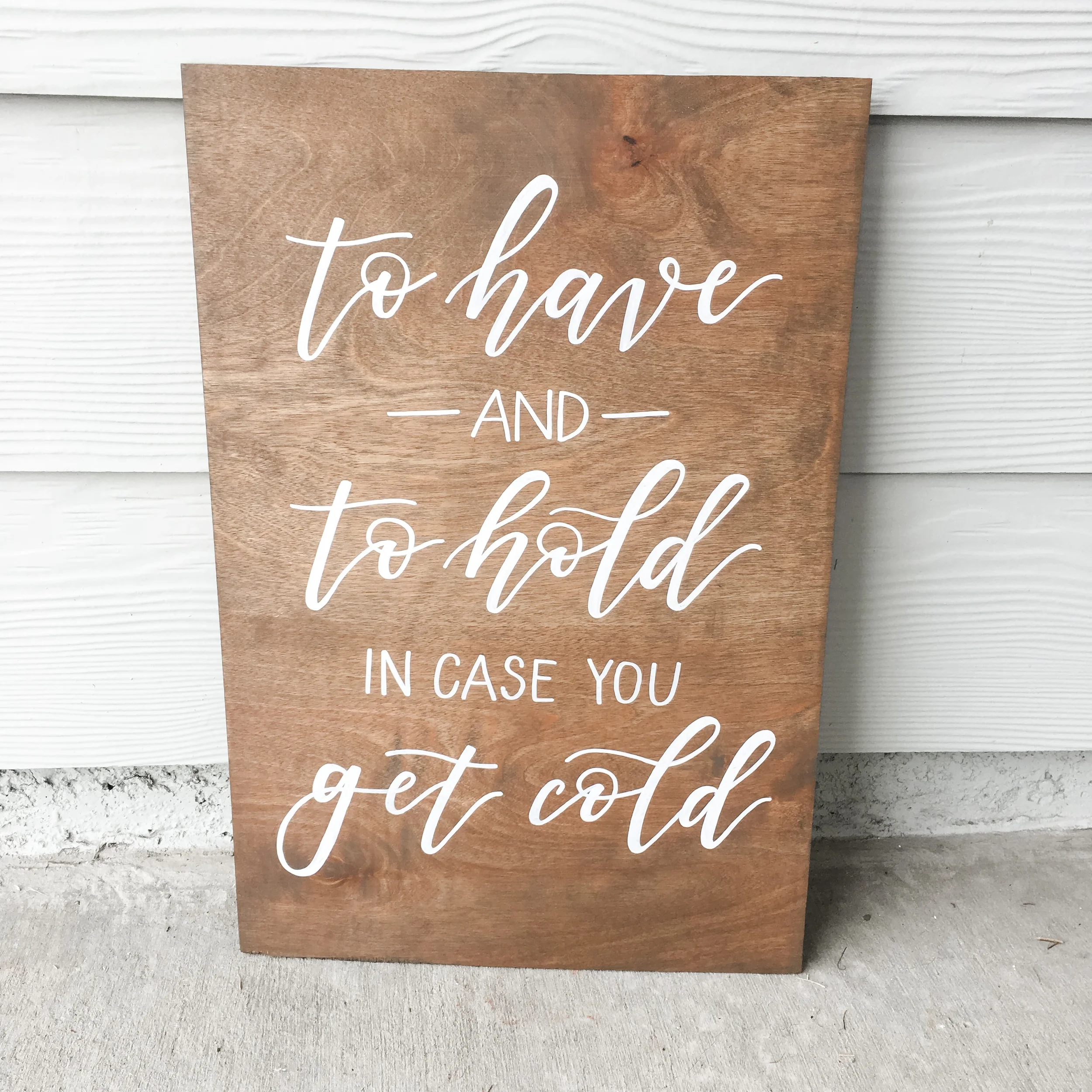 Wooden sign hand-lettered in white script reading "To have and to hold in case you get cold" for an outdoor wedding blanket station.
