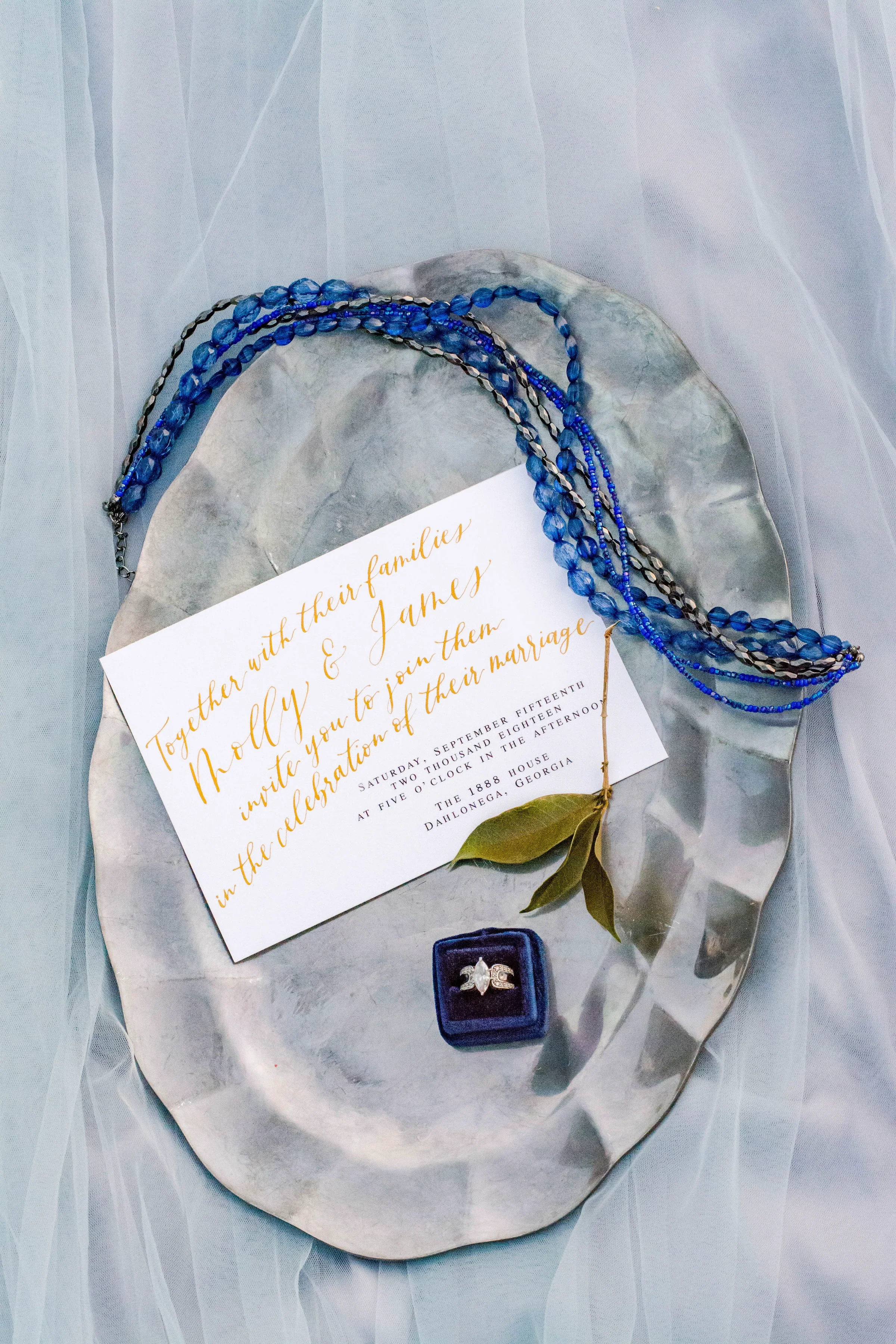 White wedding invitation with gold calligraphy resting on a decorative oval tray with blue jewelry and a velvet ring box.
