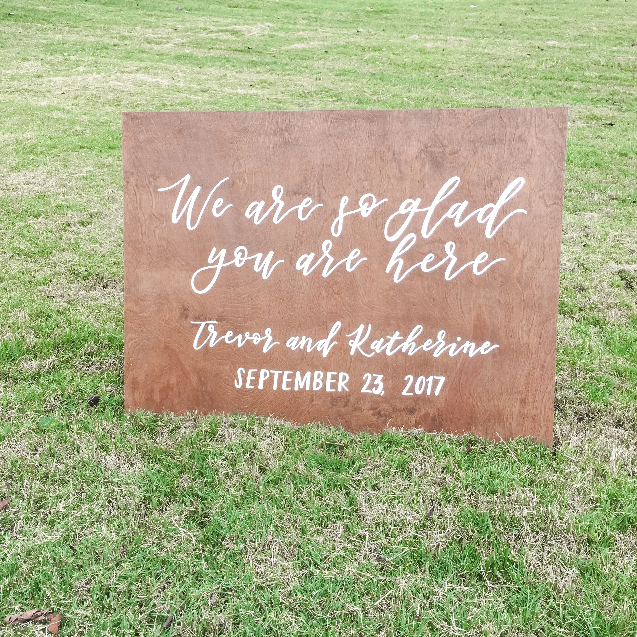 Wood wedding welcome sign that reads "we are so glad you are here" with the couples names and wedding date in white hand-lettering.