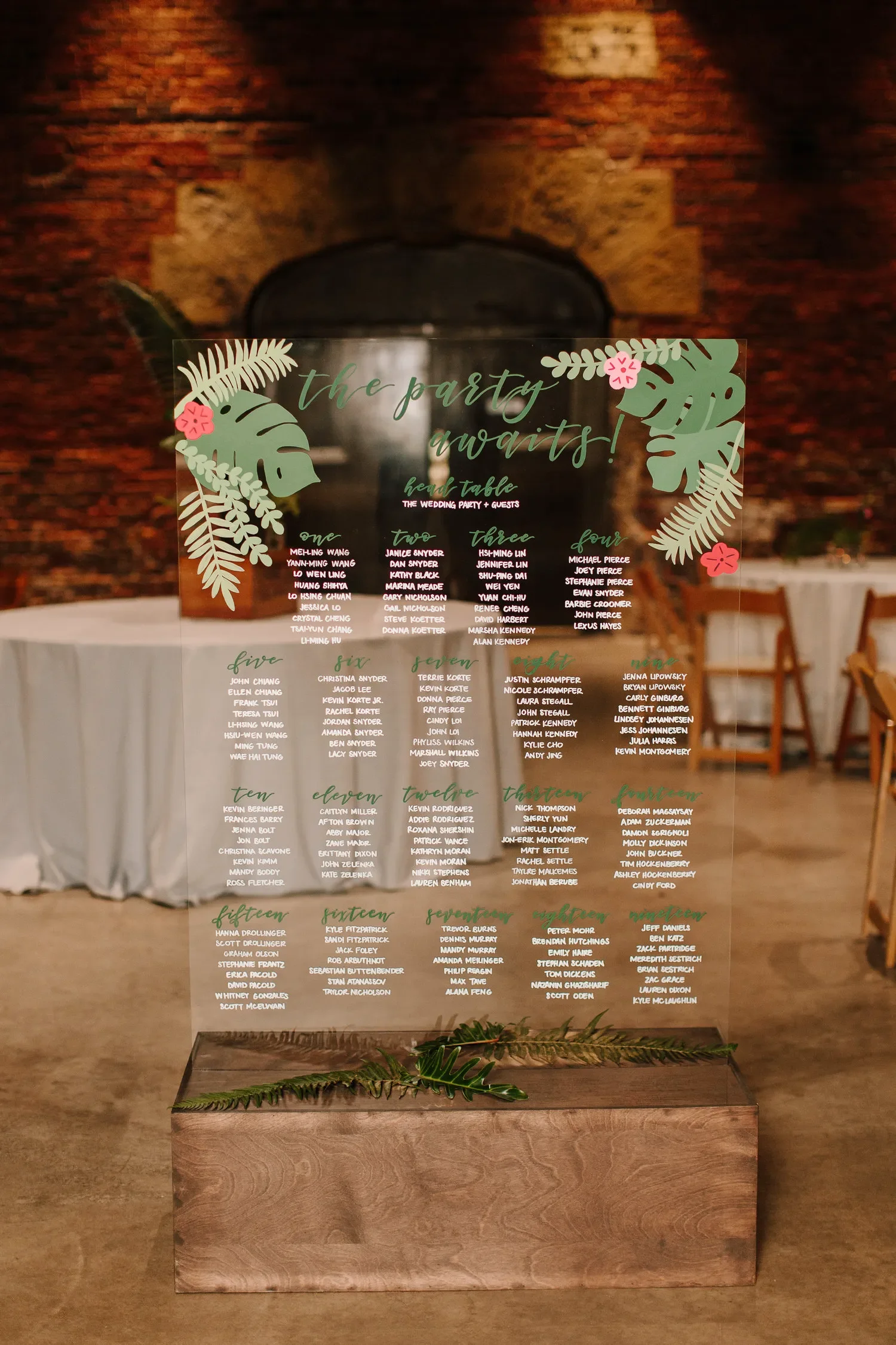 Large clear acrylic wedding seating chart with white ink font sitting on a dark wooden block.