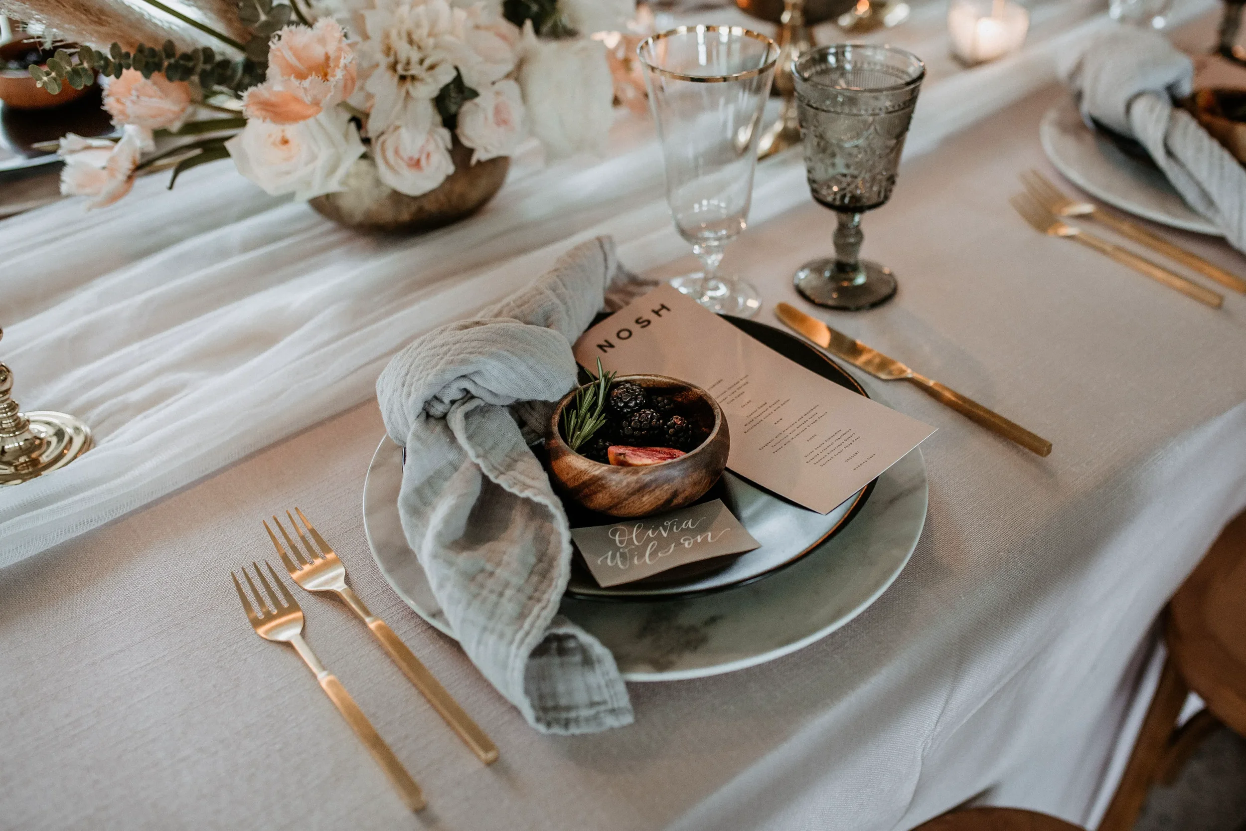 Elevate your reception dining experience with a texturally rich, moody tablescape design. This modern plate setting combines a minimalist blush menu card with a translucent clear place card written in striking white ink calligraphy, framed elegantly 