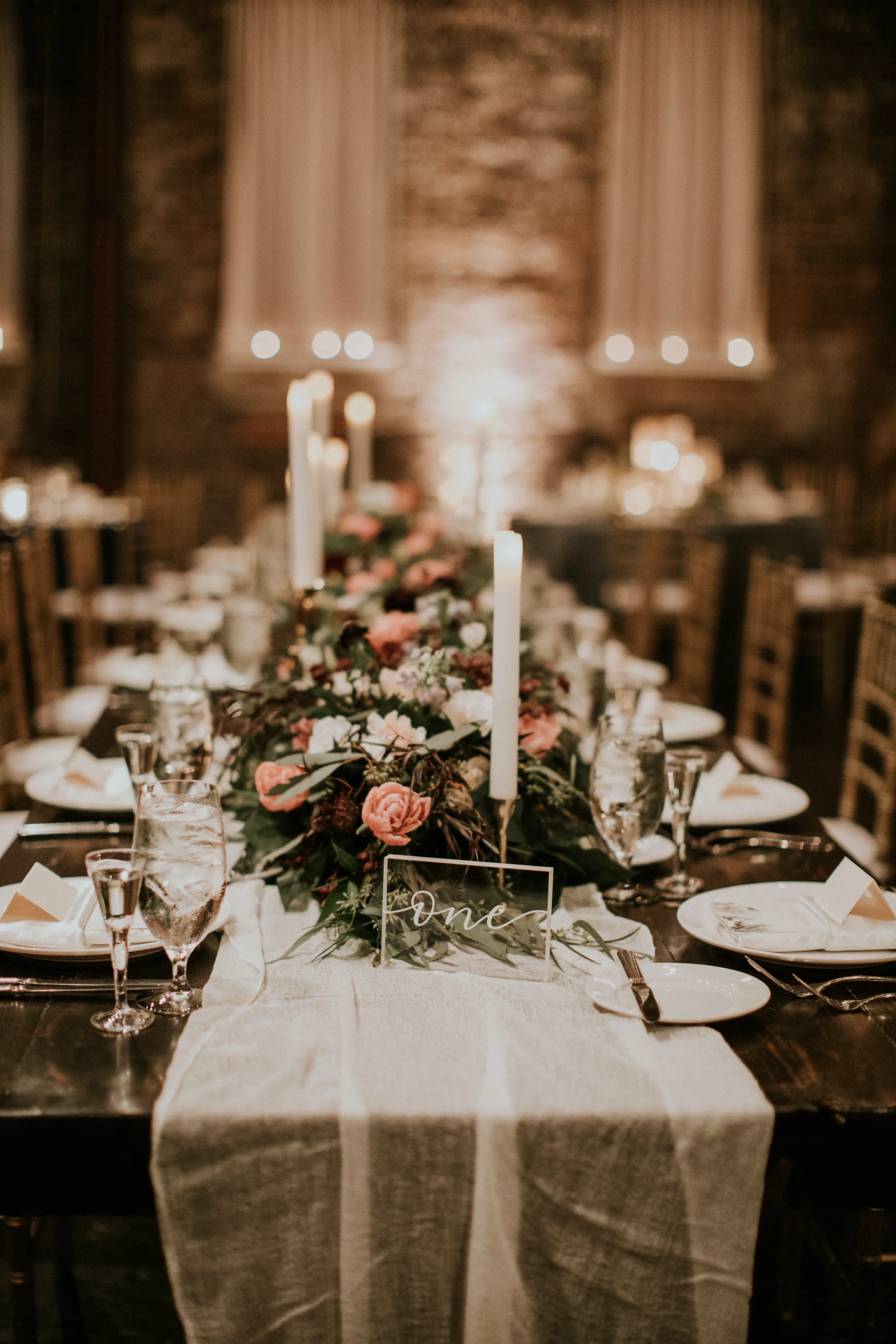 Clear acrylic block wedding table number with white script calligraphy reading "one" placed on a wooden farm table next to a low floral centerpiece, white gauze runner, and tall taper candles.