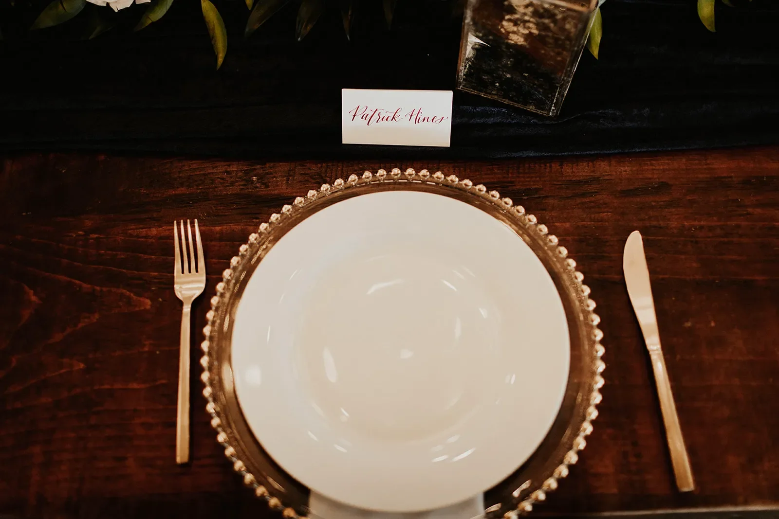 Infuse classic fine-art luxury into your tabletop presentation with traditional hand-lettered wedding place cards. This clean white card features a custom name written in rich burgundy script calligraphy, resting delicately on a gold beaded charger p