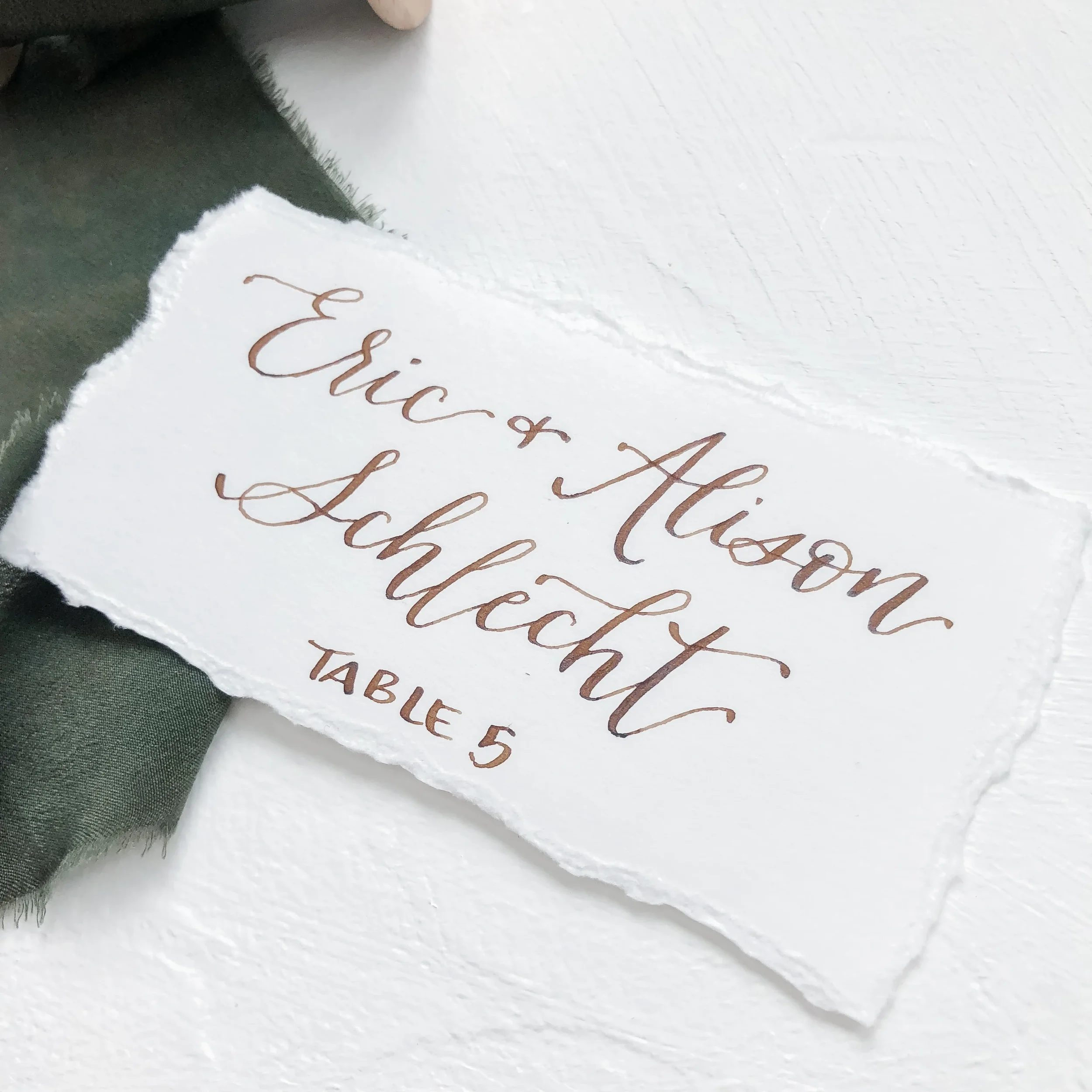 Minimalist white flat wedding escort card with walnut brown calligraphy script and torn edges.