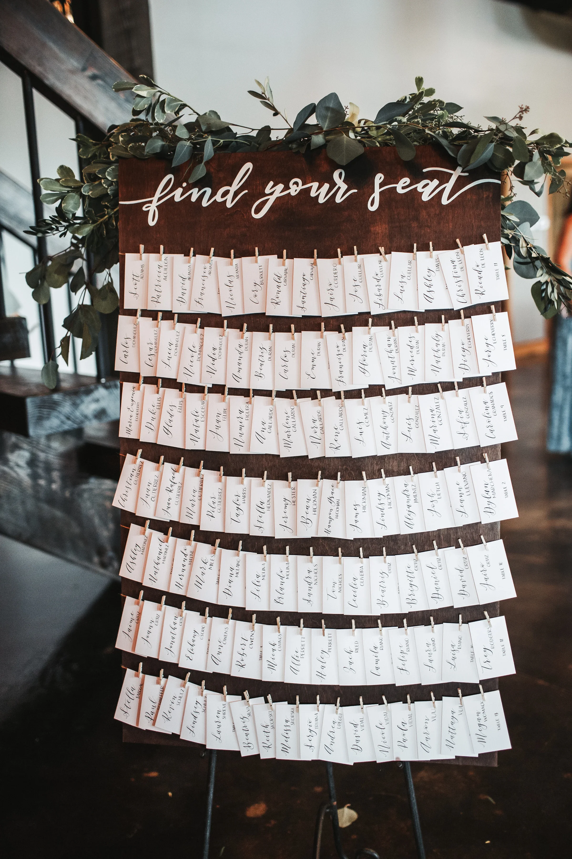 Add interactive charm to your reception entrance with a rustic clothesline escort card display. Featuring a dark stained wooden board topped with lush greenery and white script lettering reading "Find Your Seat," this setup lets guests easily find an