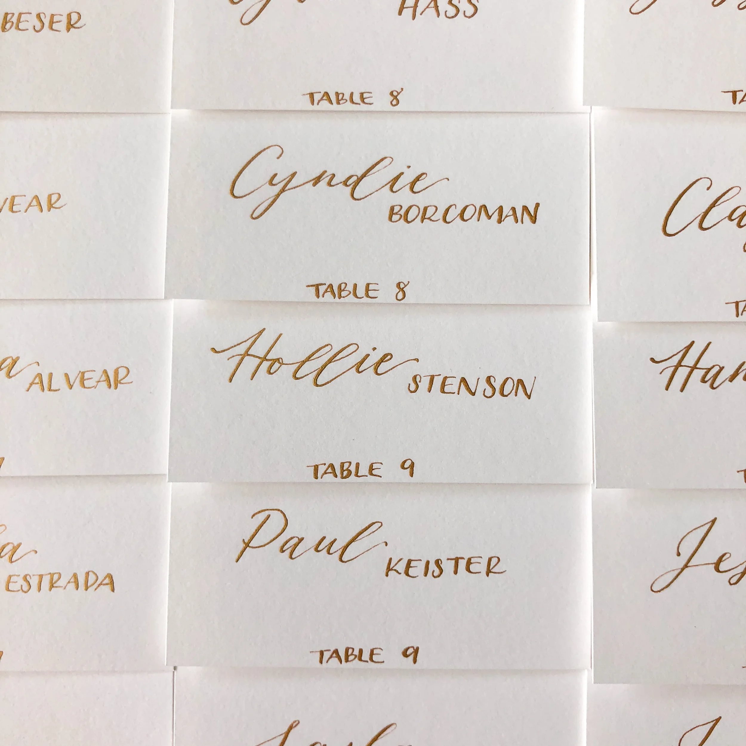Close-up of clean white paper seating cards featuring custom gold hand lettering for guest table assignments.
