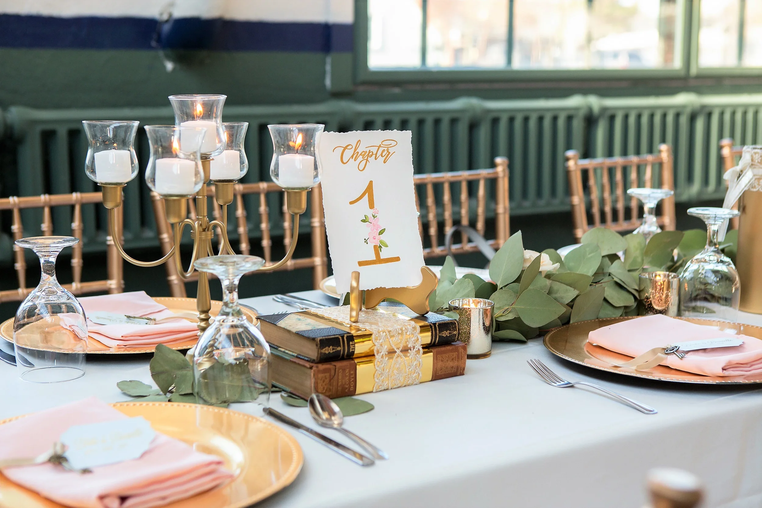 Romantic floral-illustrated wedding table sign placed on a gold easel over stacked antique books.