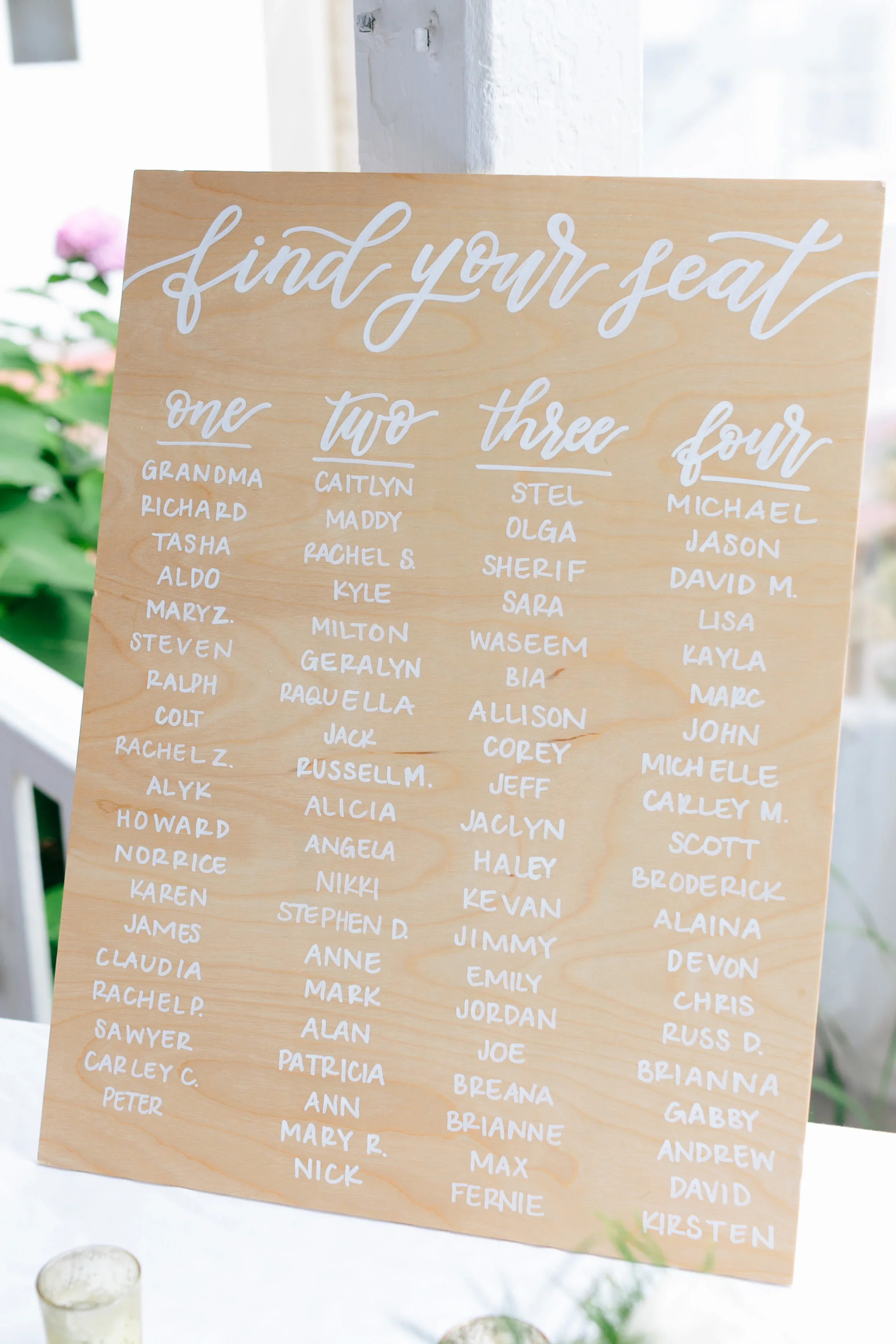 Light wood plywood seating chart sign with "Find Your Seat" hand-lettered in white cursive calligraphy on a white tablecloth with votive candles.