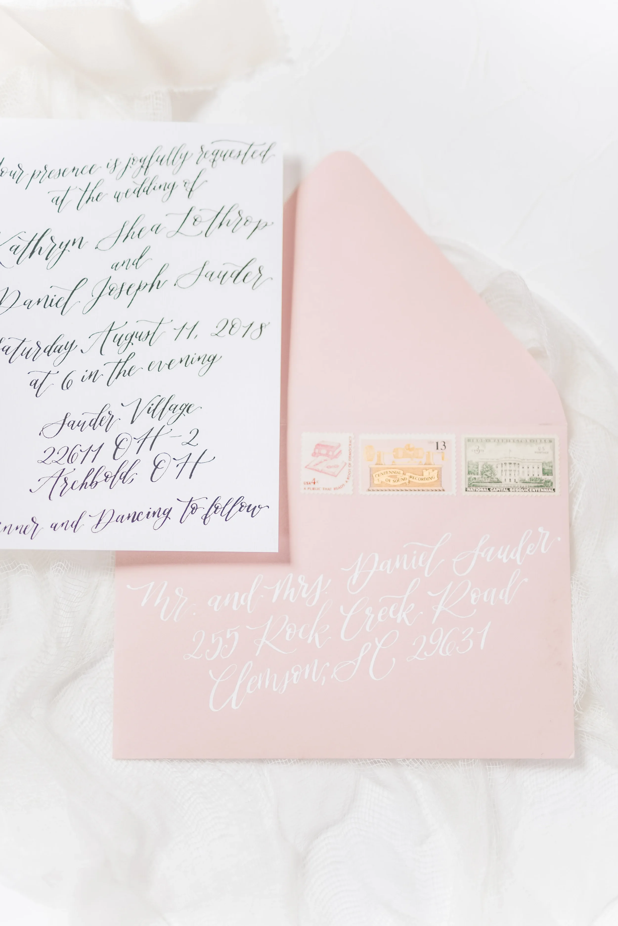 Blush pink wedding envelope addressed in white ink calligraphy with vintage stamps alongside a formal script invitation card.