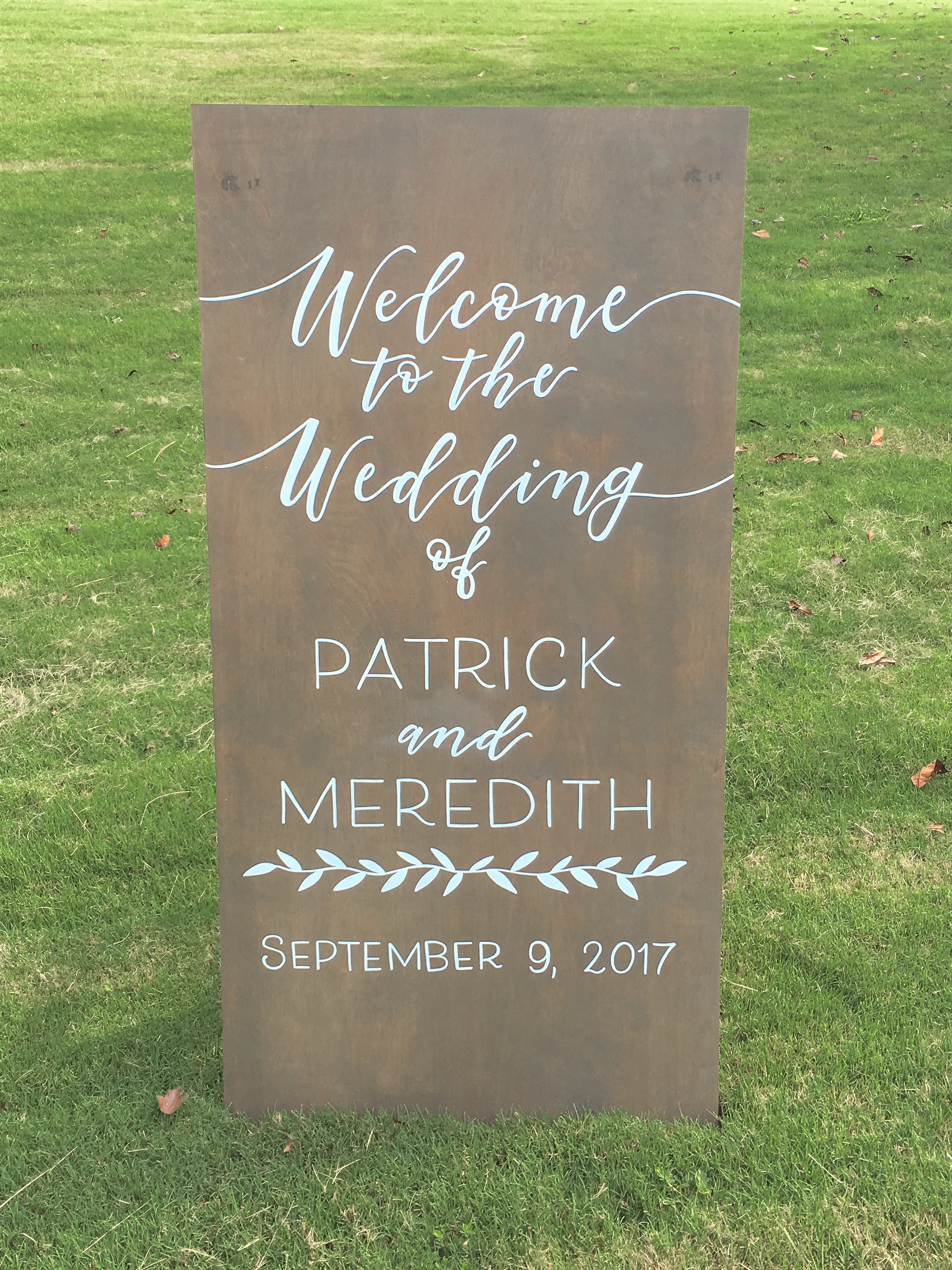 Tall weathered grey-stained wood welcome sign reading "Welcome to the Wedding of Patrick and Meredith" with botanical branch accents.