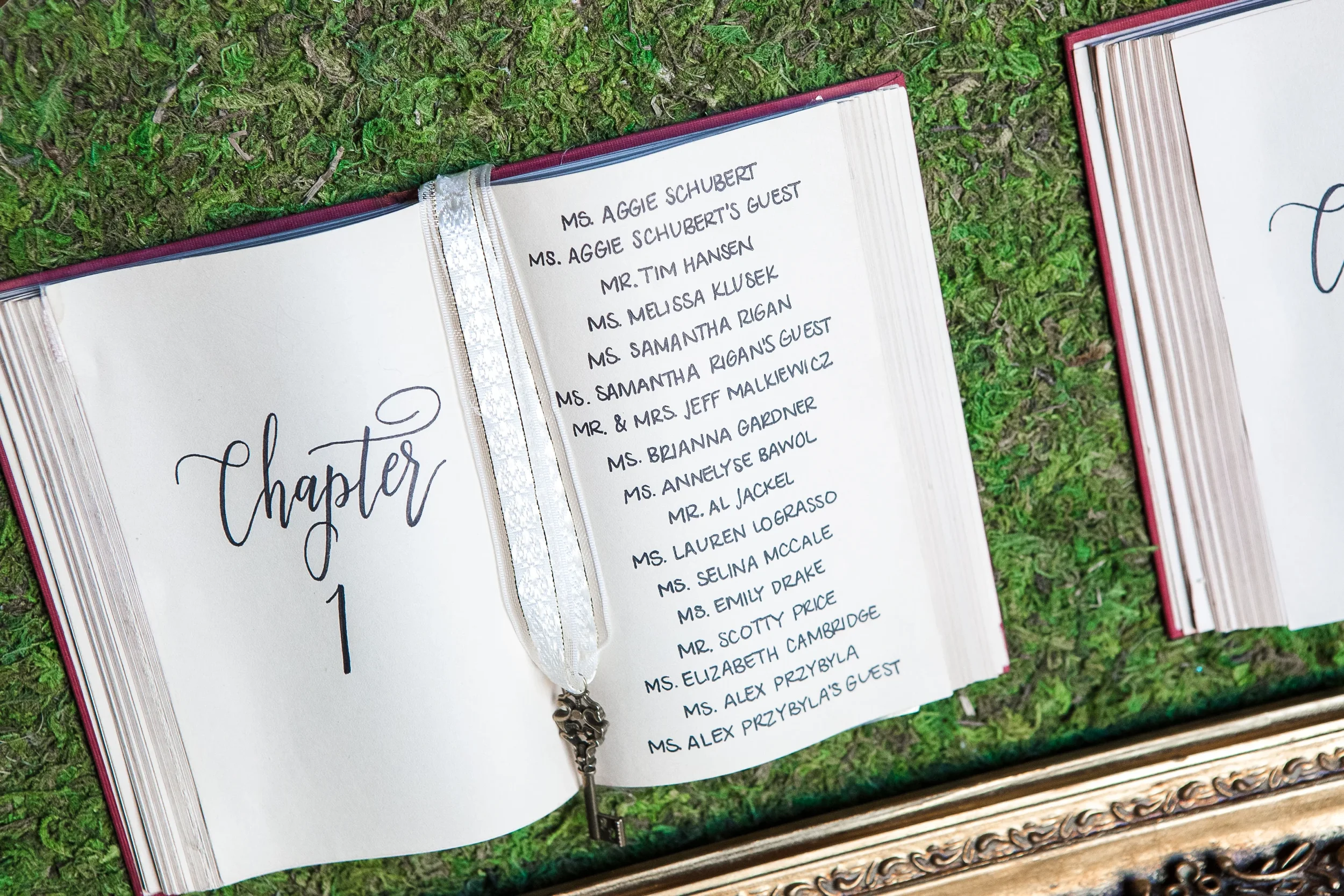 Bring fairytale romance to your reception with a storybook open-book seating chart display. Framed in a opulent vintage gold baroque frame against a vibrant moss backing, each open hardcover book features calligraphed "Chapter" table numbers, hand-wr