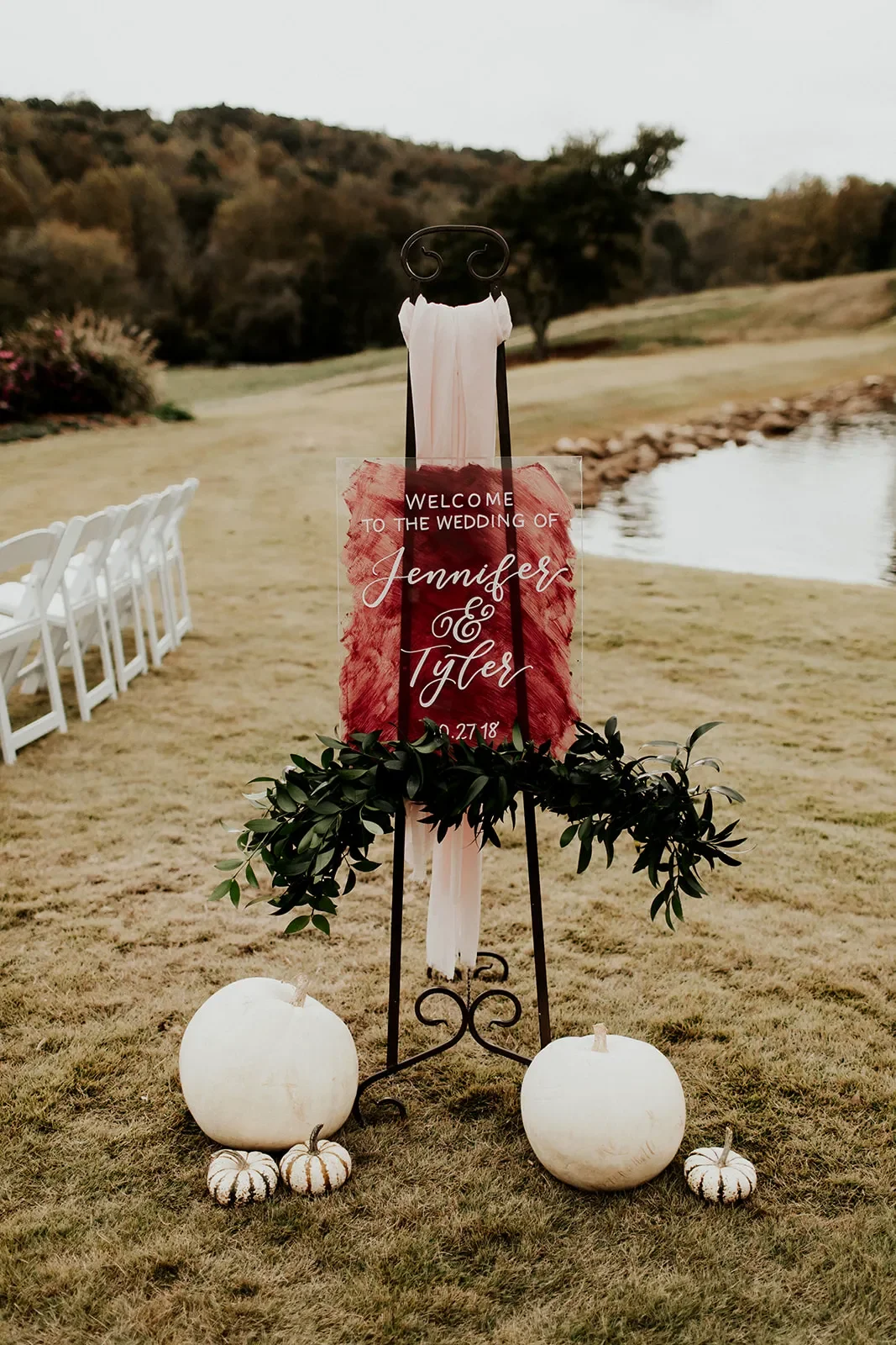 Burgundy painted-back acrylic wedding welcome sign on a black easel with white pumpkins.
