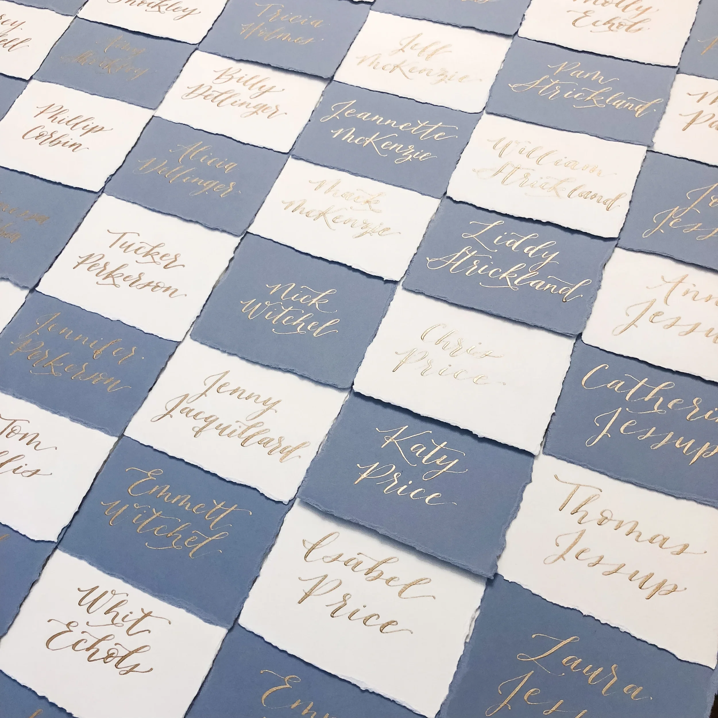 High-angle view of handmade paper escort cards arranged in a checkerboard pattern with gold ink hand lettering.High-angle view of handmade paper escort cards arranged in a checkerboard pattern with gold ink hand lettering.