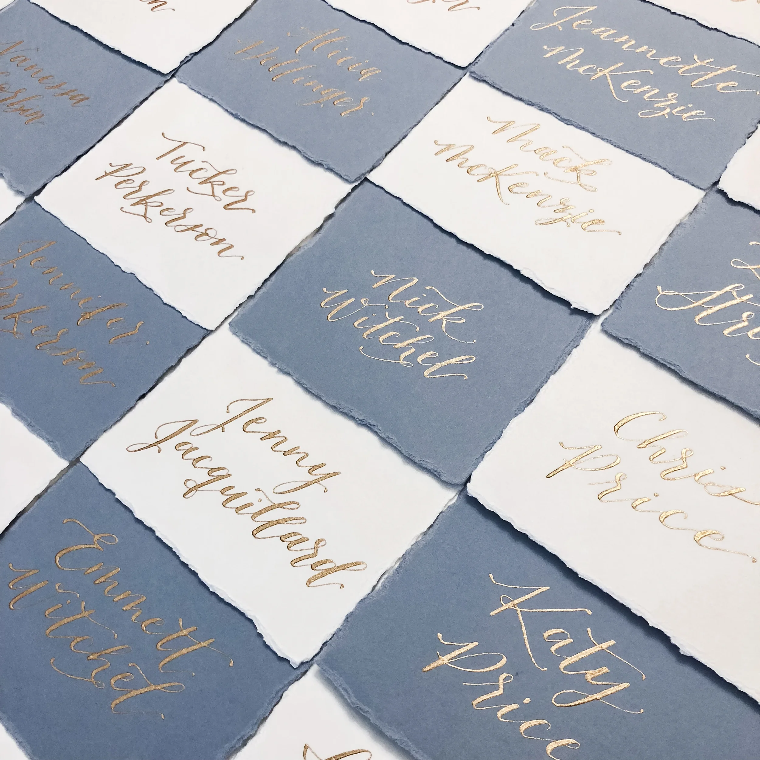 Elevate your reception seating display with alternating dusty blue and ivory deckled-edge place cards. Hand-lettered in shimmering metallic gold calligraphy script, these raw-edge handmade paper escort cards create an eye-catching, sophisticated grid