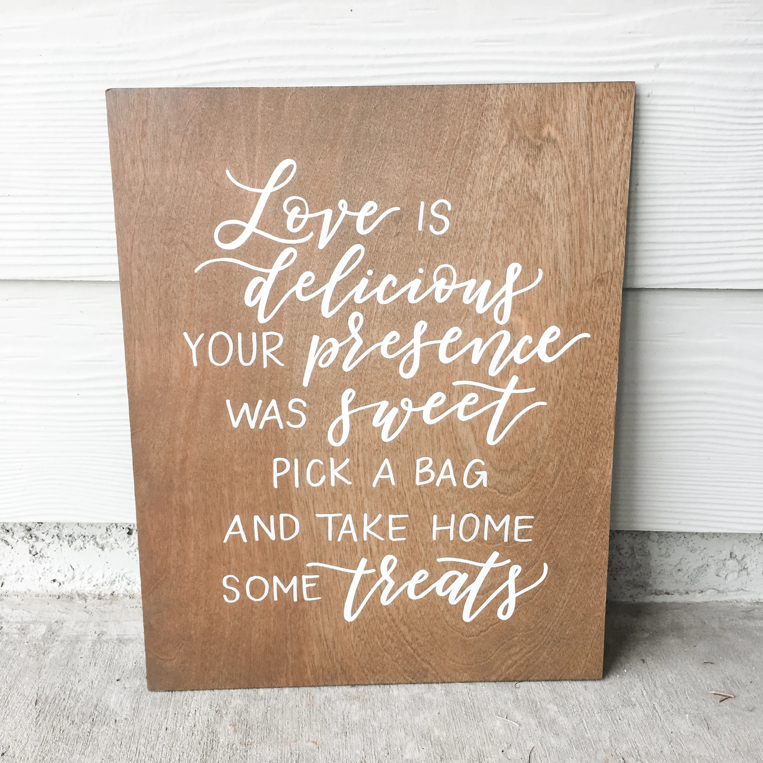 Stained wood sign reading "Love is delicious your presence was sweet pick a bag and take home some treats" in white script.