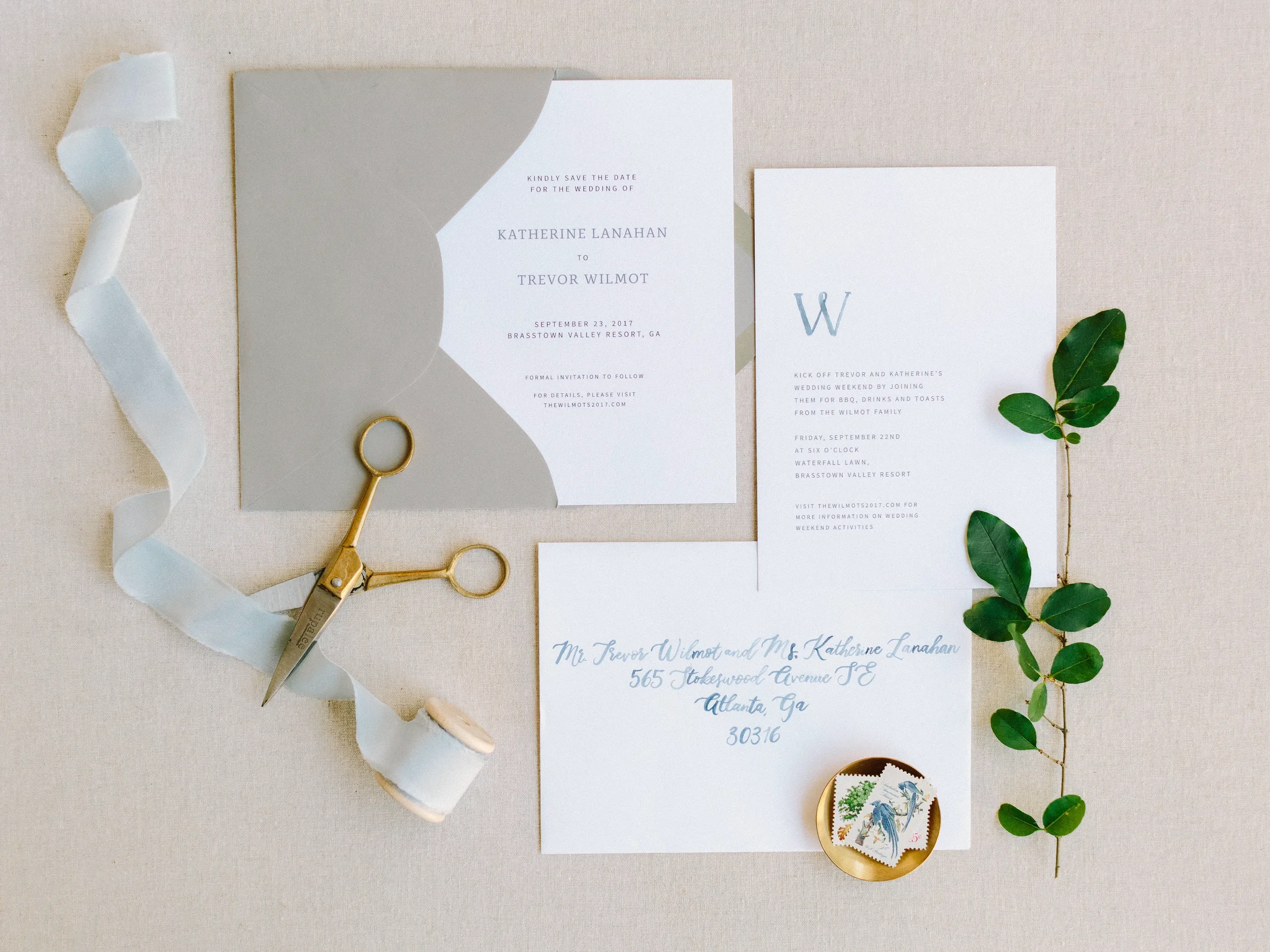 Minimalist grey and white wedding stationery with watercolor envelope addressing flat lay detailed with a green foliage stem and vintage bird postage stamps.