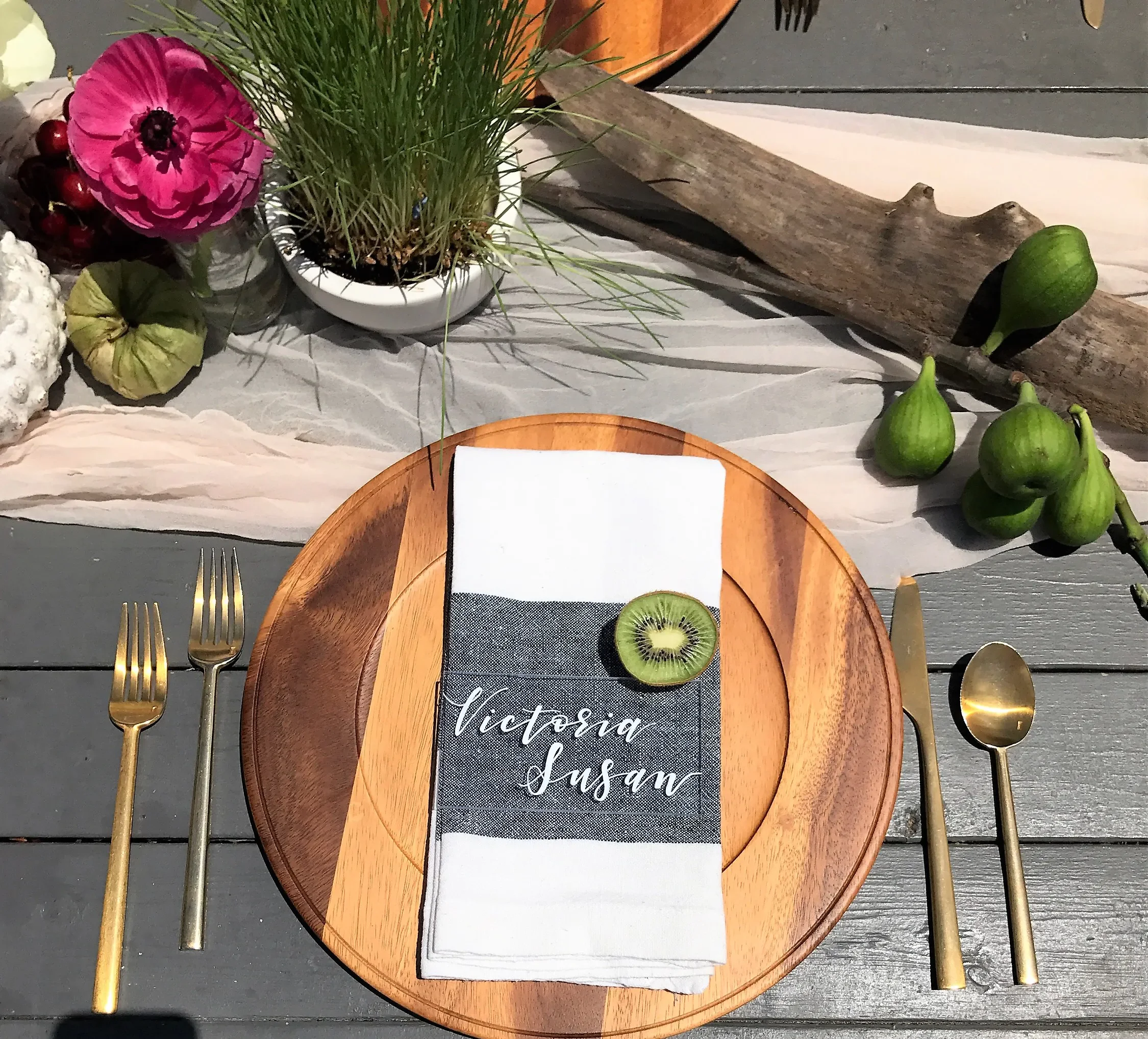 Rustic wooden charger plate with a white napkin, slate grey calligraphy menu strip, fresh kiwi slice, and gold flatware.