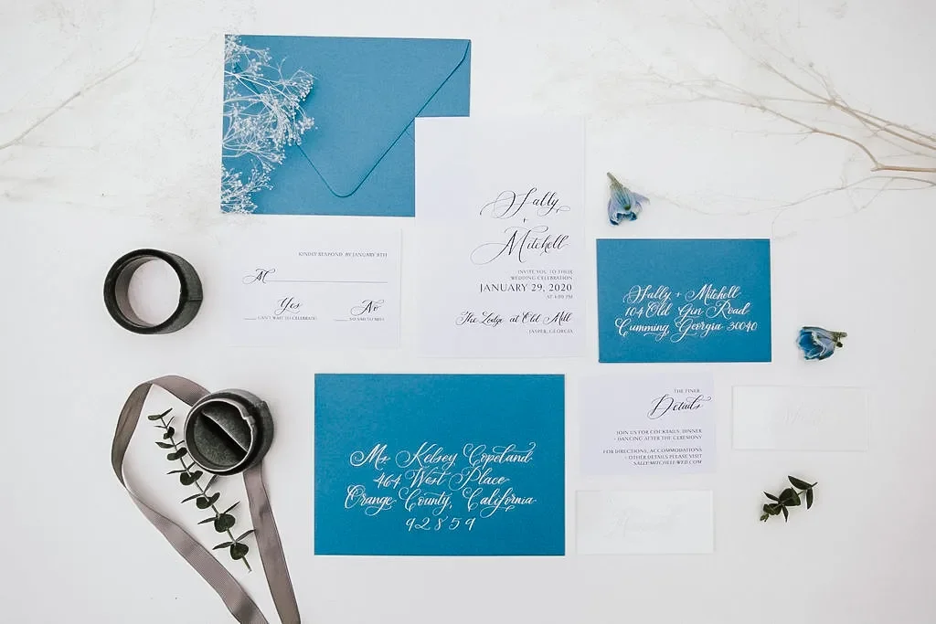 Monochromatic ocean blue and dusty teal wedding invitation suite styled with a ribbon spool and dried florals.