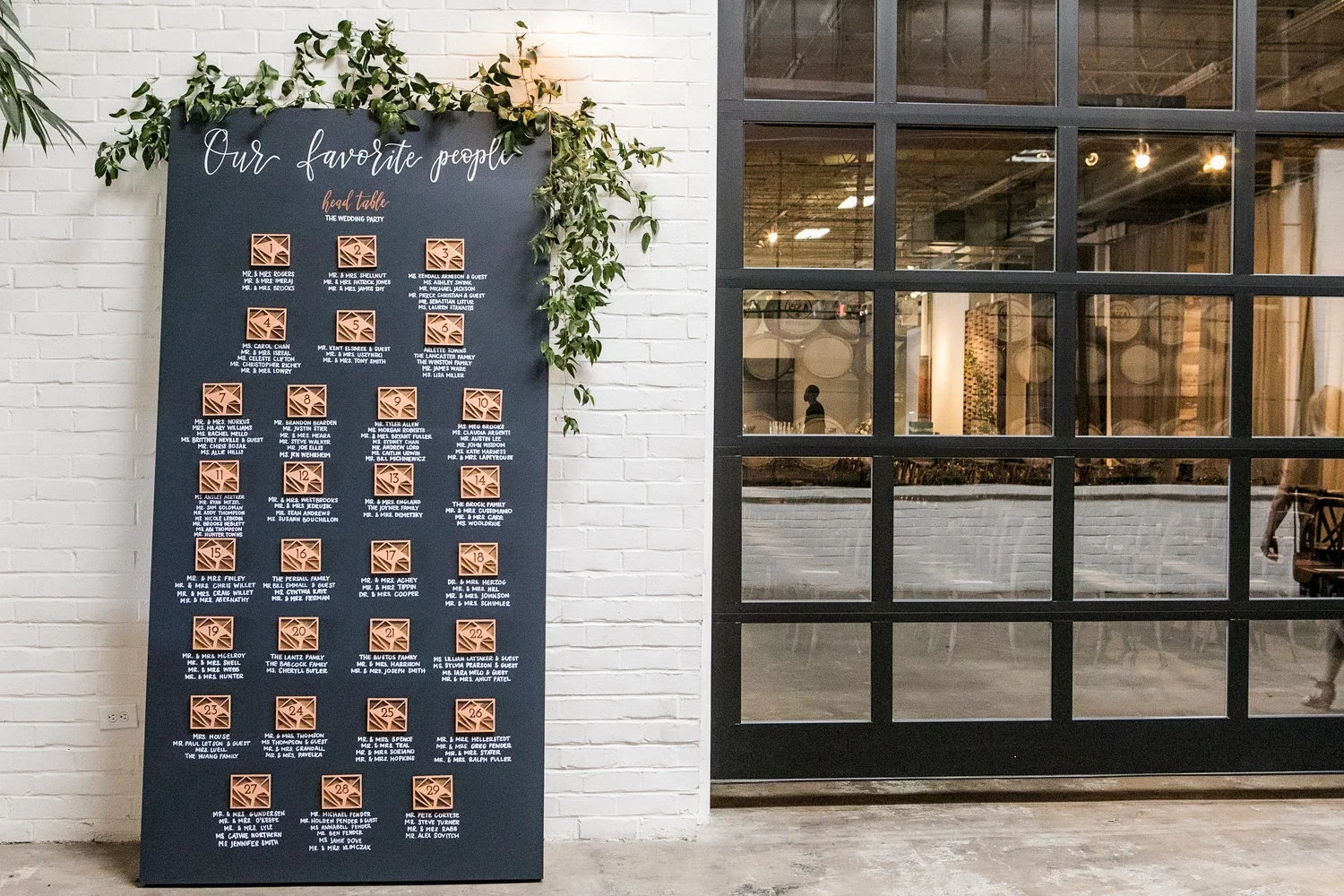 Create high-contrast drama at your venue entrance with a large black vertical seating chart. Adorned with organic greenery overhead and titled "Our favorite people," this striking dark board stands out sharply against industrial white brick walls for