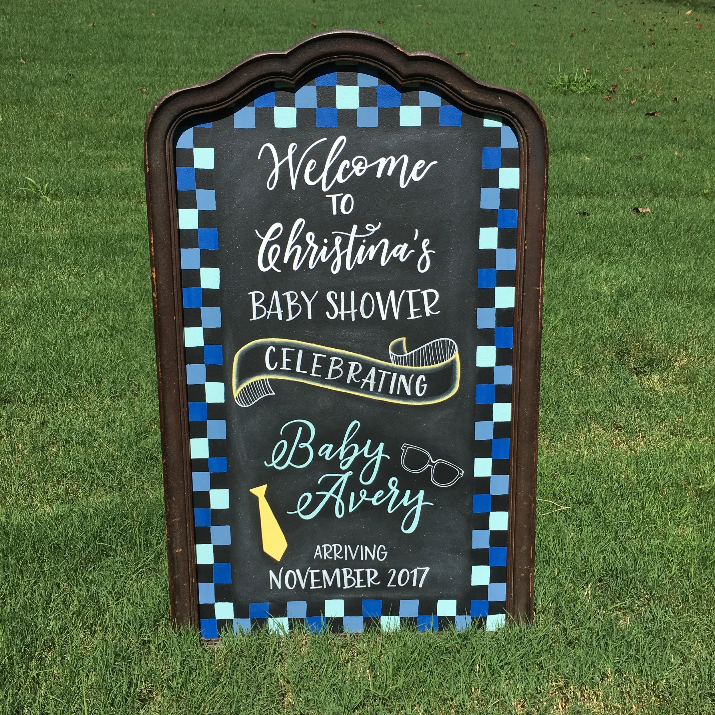 Hand-lettered baby boy shower chalkboard sign with colorful checkered border and illustrations.