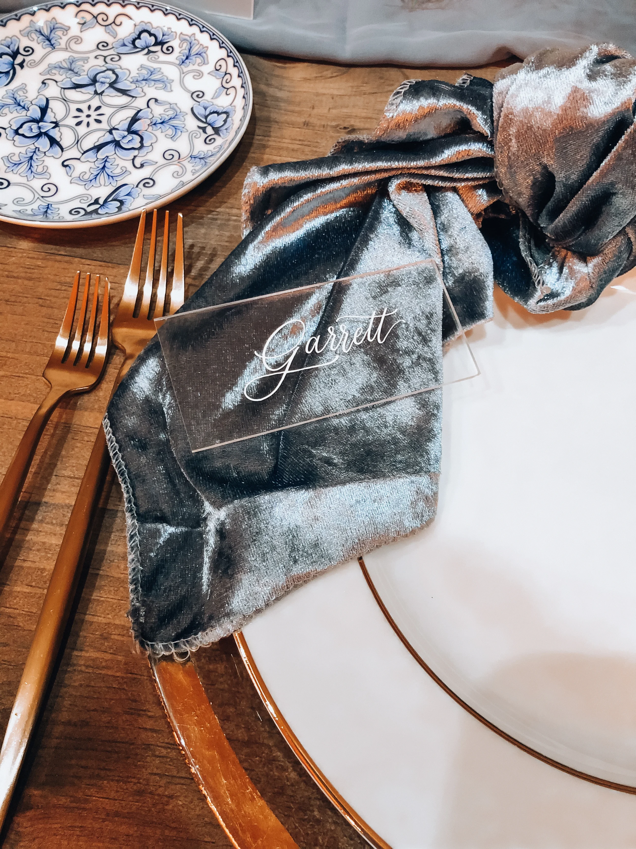 Clear acrylic place card with white script calligraphy on a plush slate blue velvet napkin.