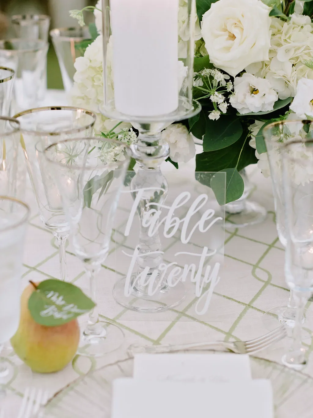 Clear acrylic table number sign with white calligraphy, styled with a pear escort card, white floral centerpiece, and elegant wedding reception table decor.