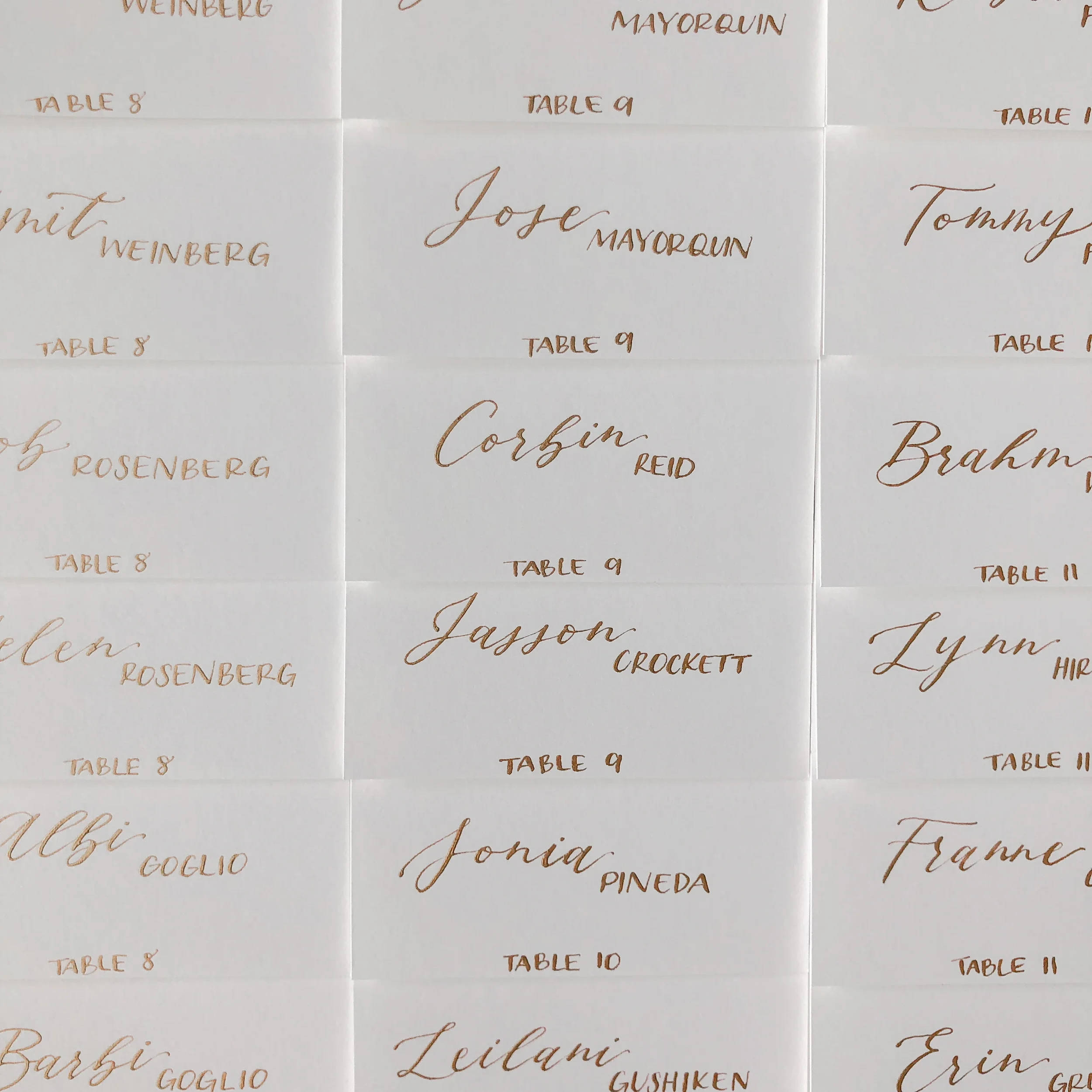 Grid layout of white wedding escort cards hand-lettered in metallic gold script calligraphy with table numbers.
