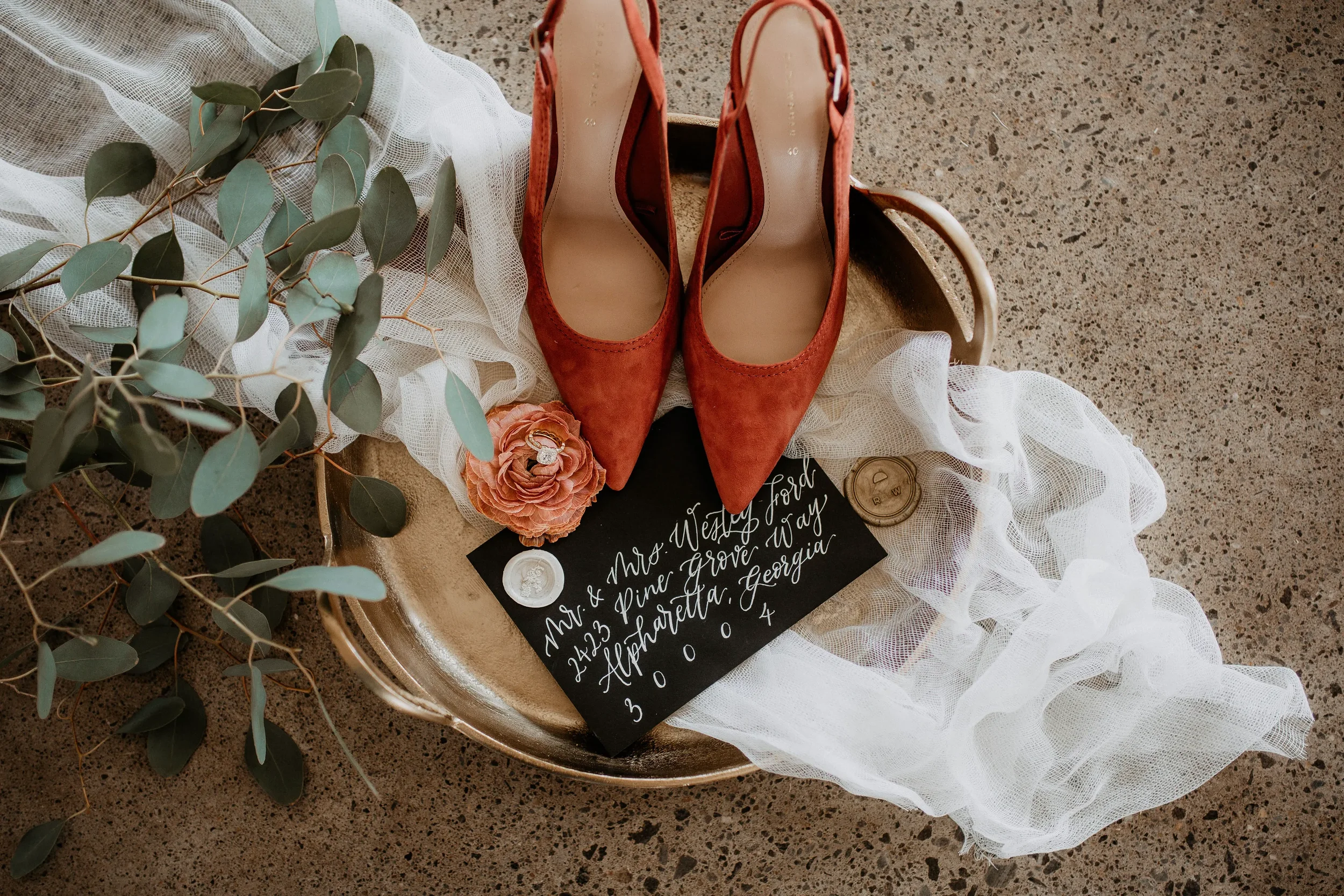 Capture the cozy, luxurious palette of a fall celebration with a curated morning detail shot. Placed on an ornate antique brass tray, vibrant terracotta orange suede heels complement a dramatic matte black guest envelope, lettered beautifully in high