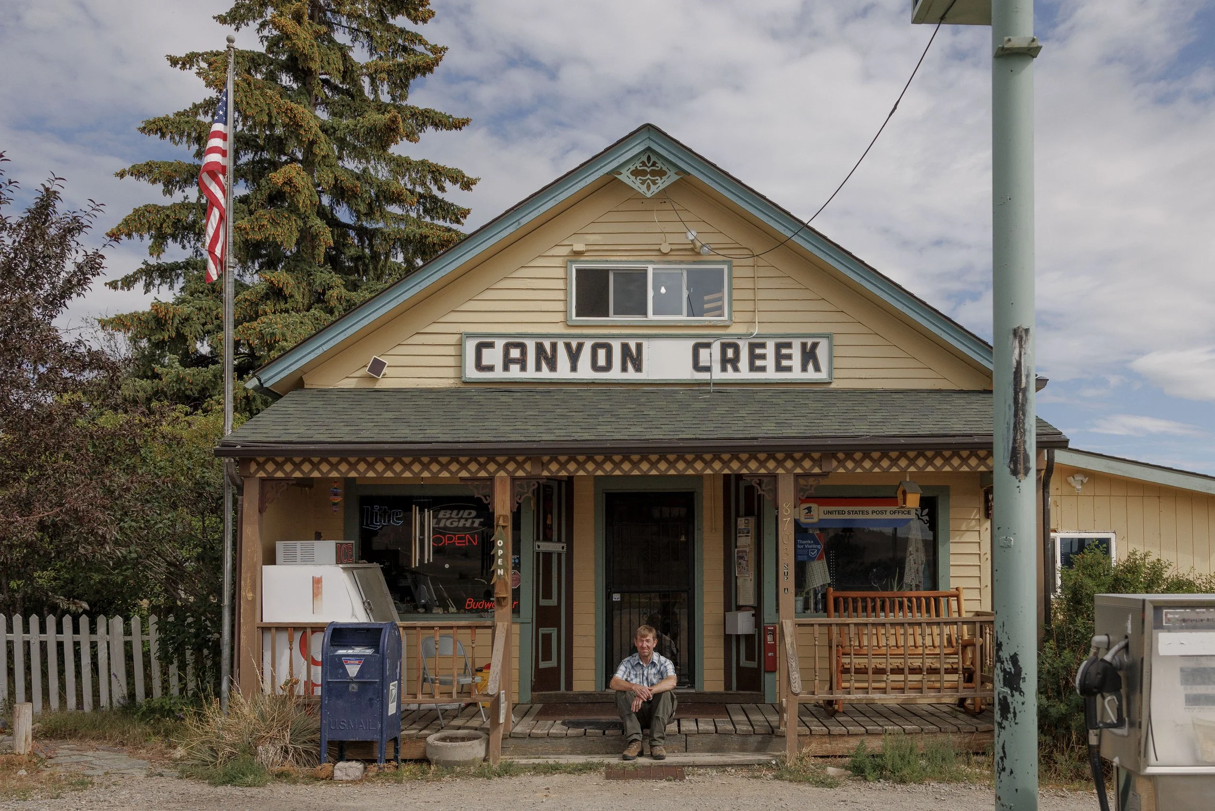 A small, yellow, wooden building with a sign that reads 'Canyon Creek' and a man sitting on the front porch. The building has a porch with a bench, a mailbox, and some signs. An American flag is flying on a pole next to the building. There are trees 