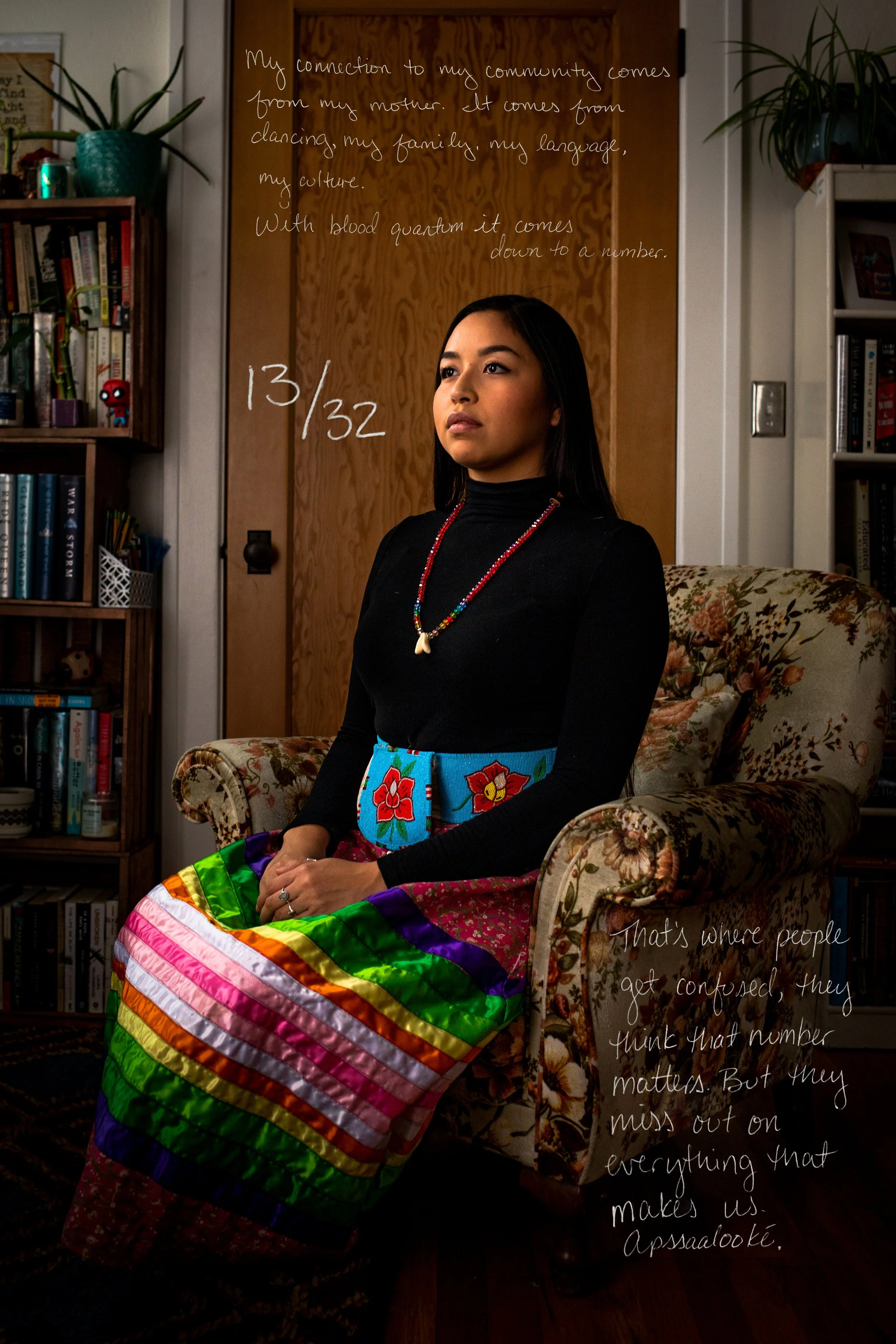 A young woman sitting on a floral upholstered armchair in a cozy room with bookshelves and potted plants. She wears a black turtleneck, colorful beaded necklace, and a vibrant rainbow-striped skirt. Behind her, a wooden door has handwritten text about cultural connection and identity, and there are personal items and decorations in the room.