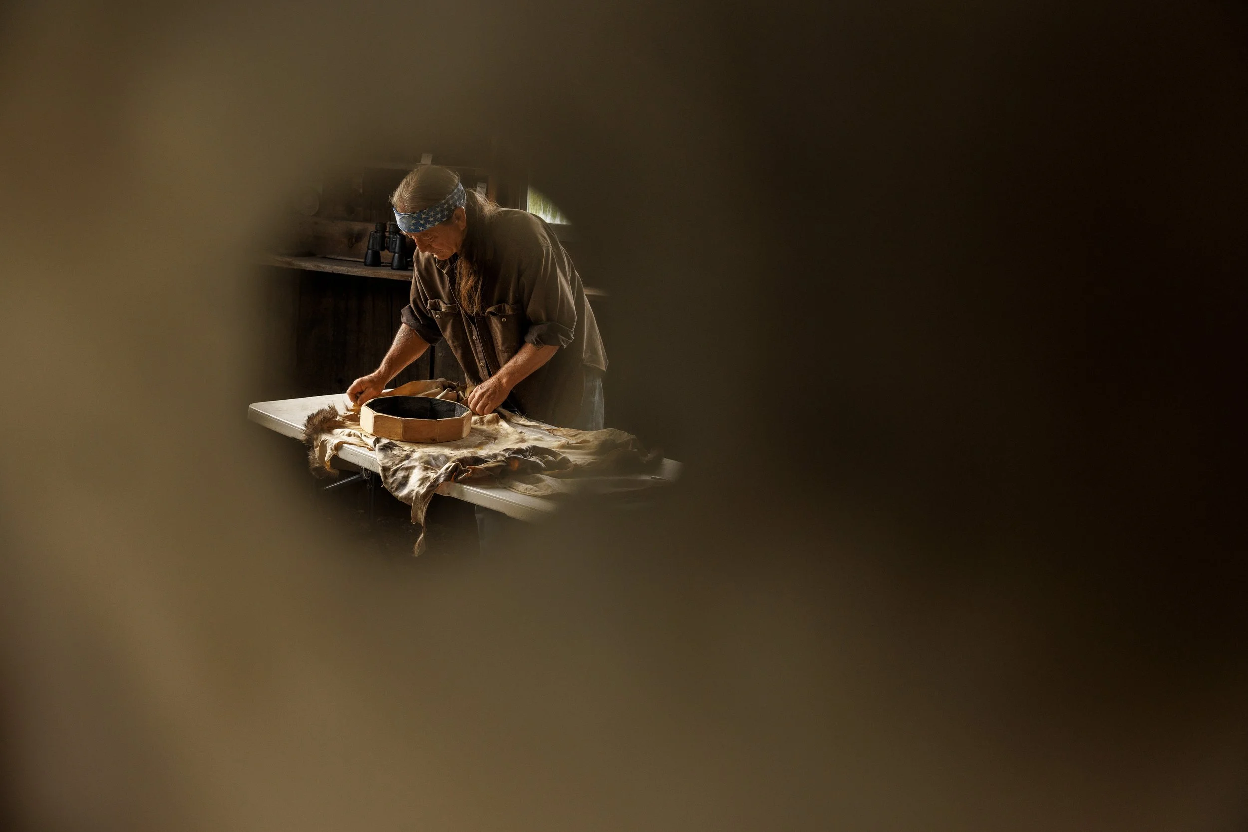 A person wearing a headband and glasses working with leather on a table, seen through a small hole.