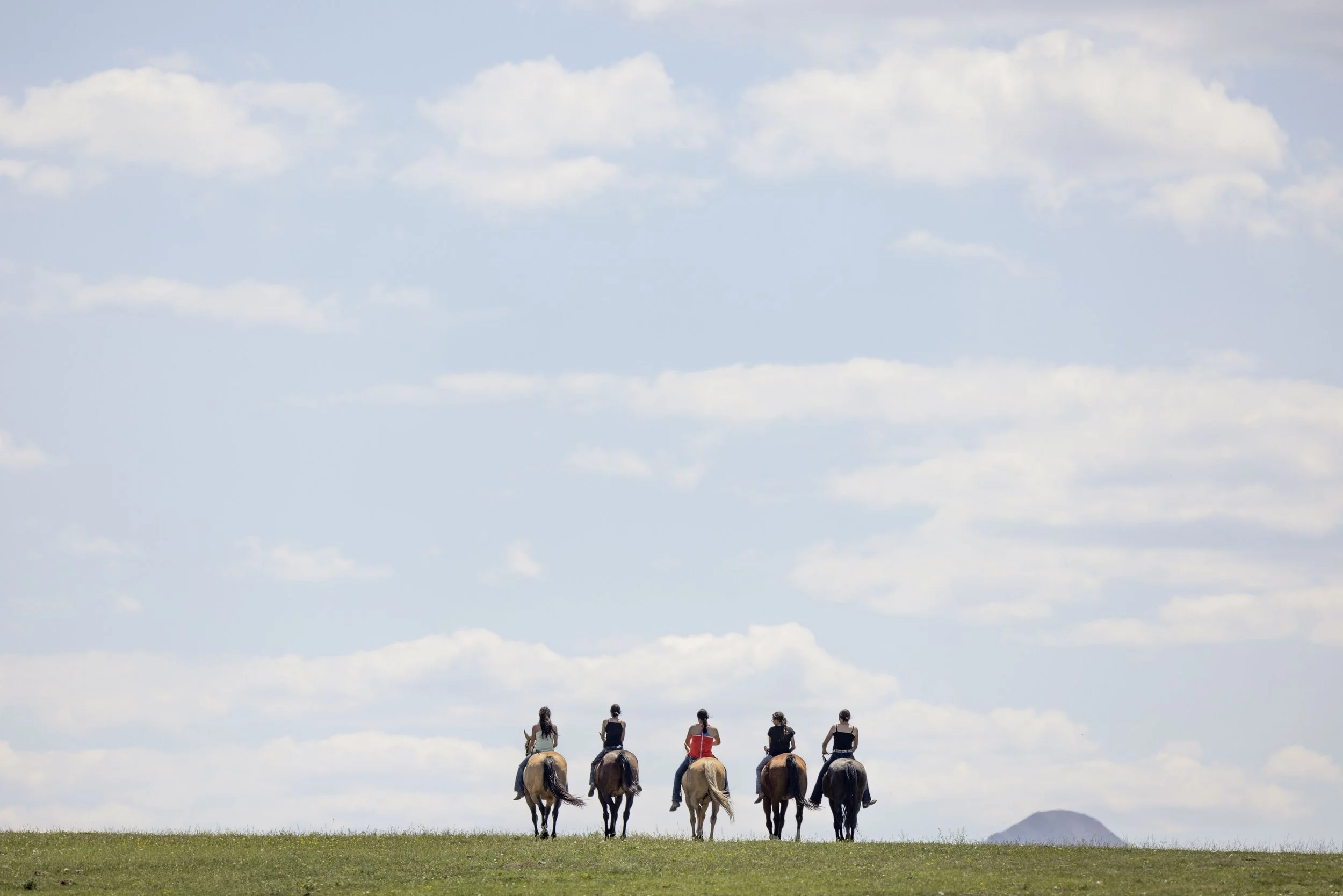 Five people riding horses in a line across a grassy field under a sky with scattered clouds, with mountains in the distance.