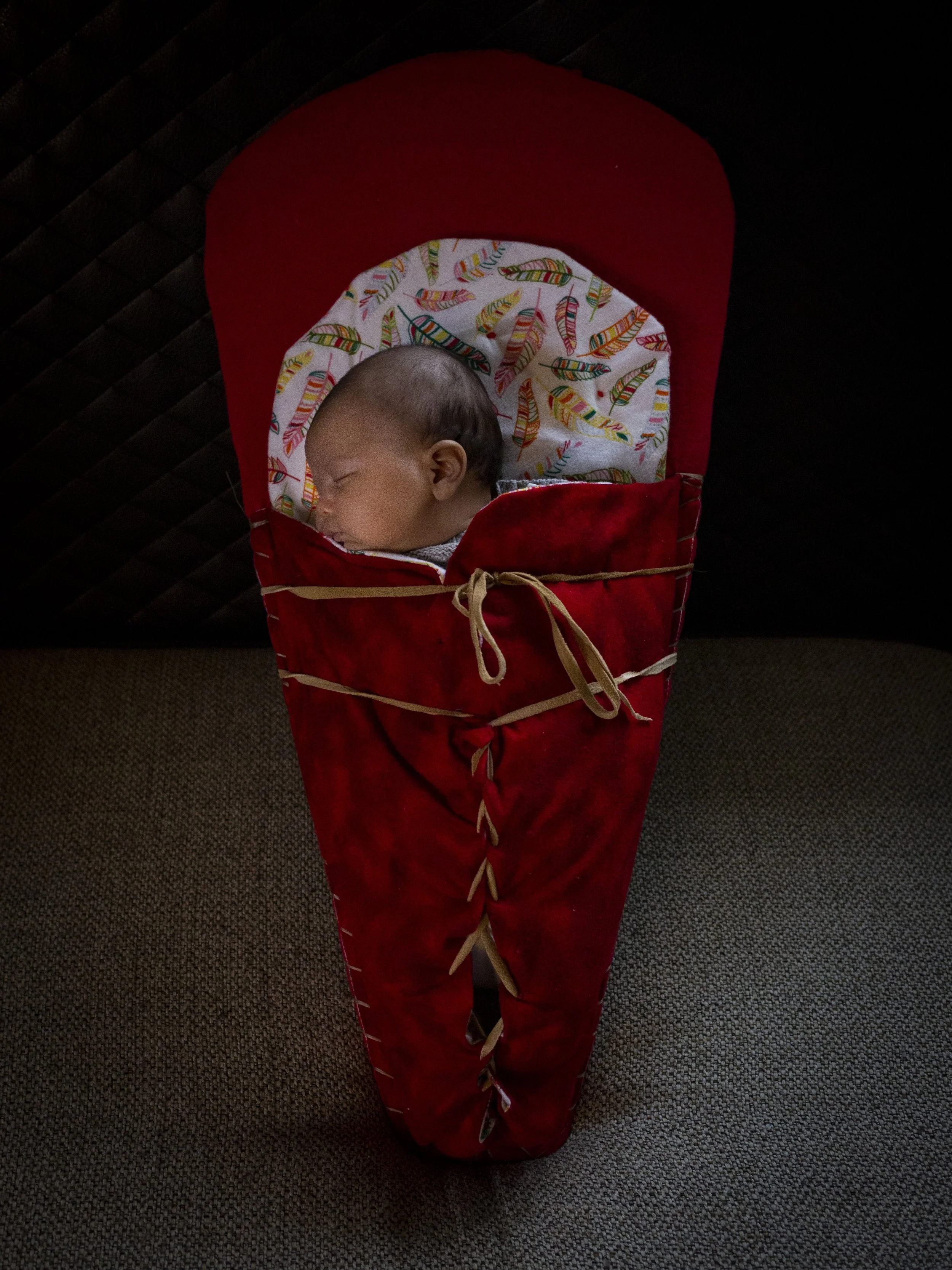 A sleeping baby wrapped in a colorful feather-patterned blanket inside a red cloth bag with beige ties.