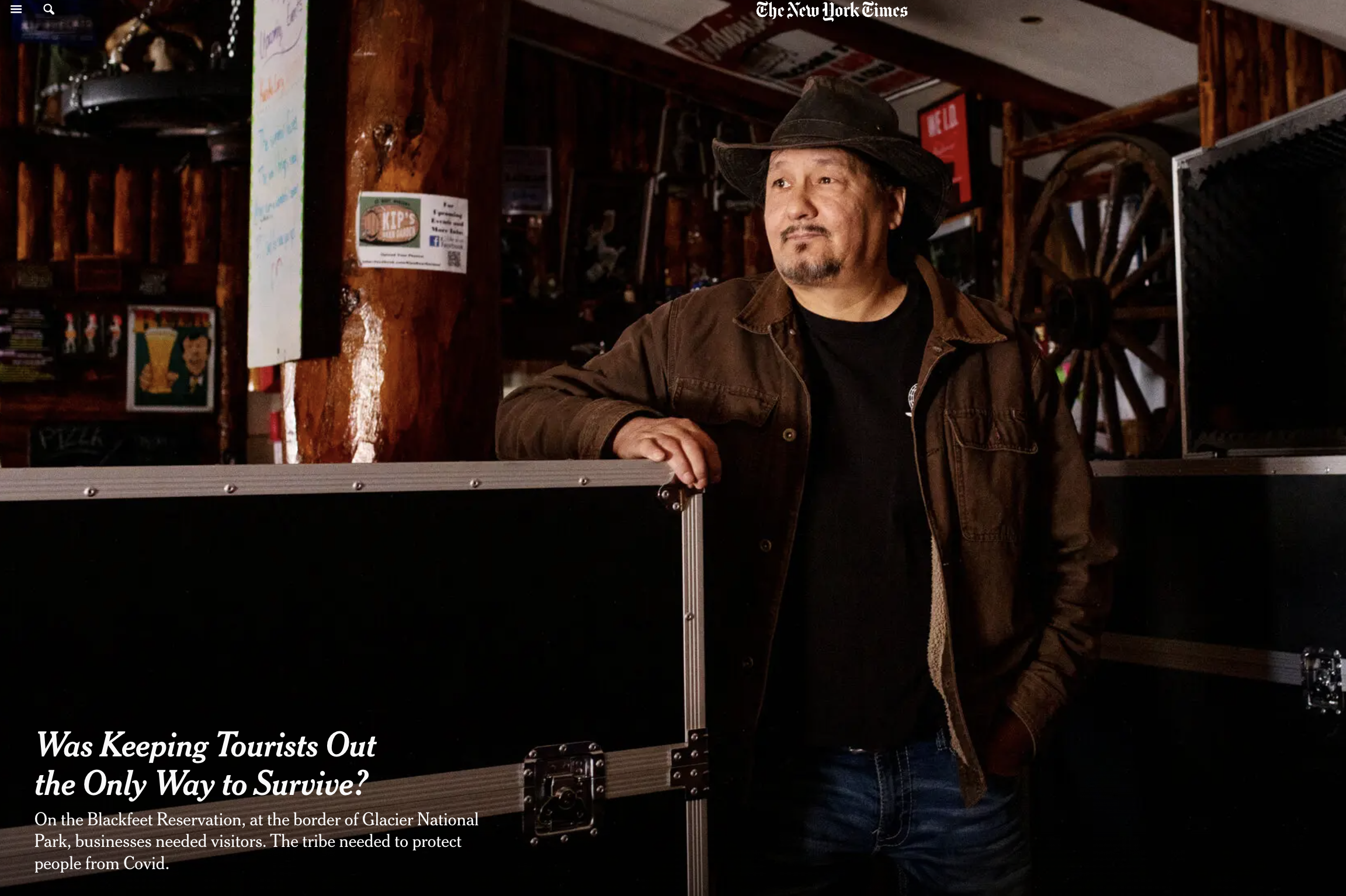 A man wearing a black hat and brown jacket standing indoors looking to his left. The background features wooden walls, a large wooden wagon wheel, and various signs and posters, including a New York Times logo and a sign about Blackfeet reservation. There is also a partially visible sign near the man reading "Was keeping tourists out the only way to survive?".