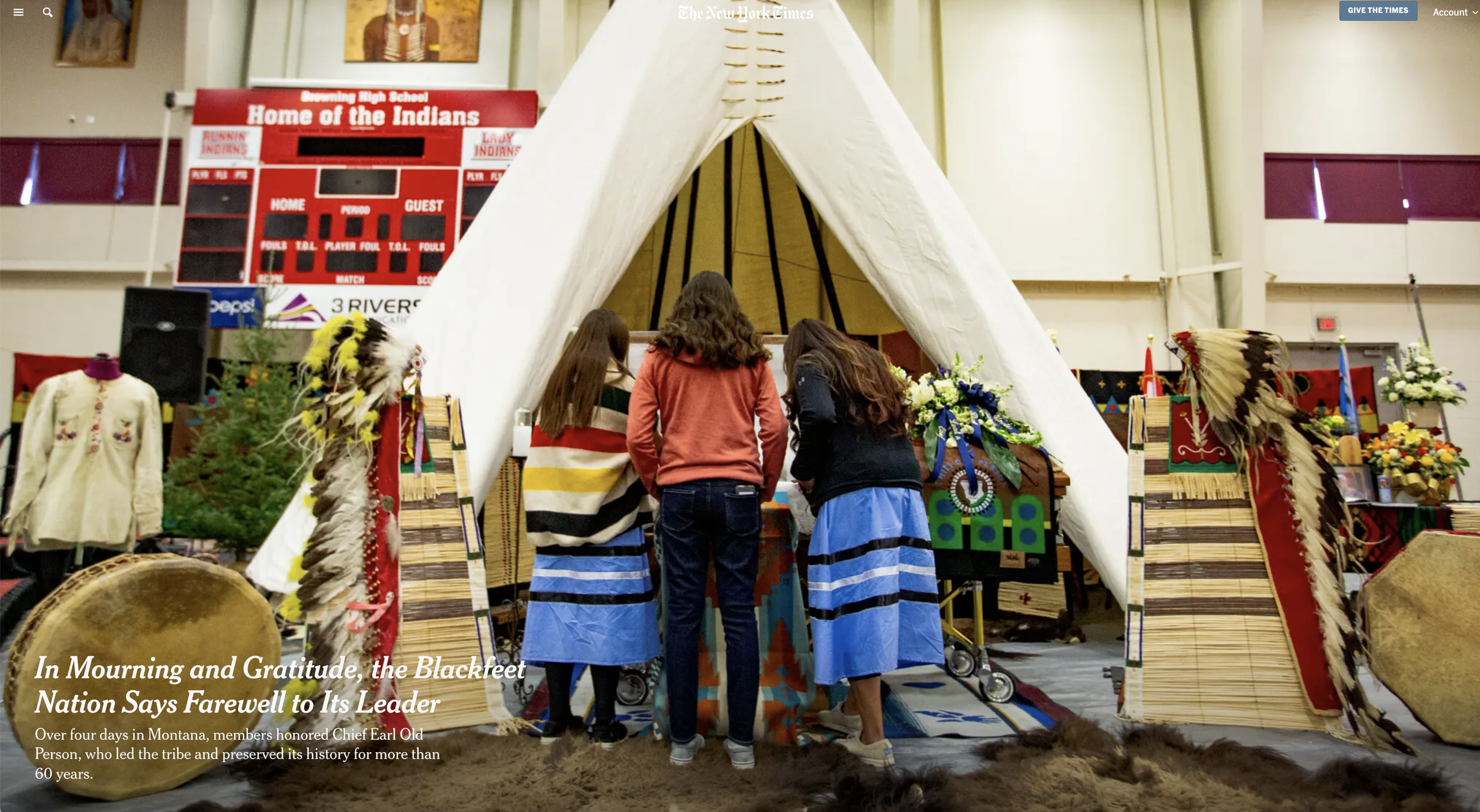 Three women are praying or paying respects inside a white tipi decorated with traditional Native American items and feathers. The background includes a scoreboard and banners, indicating the setting is an indoor arena or gymnasium.