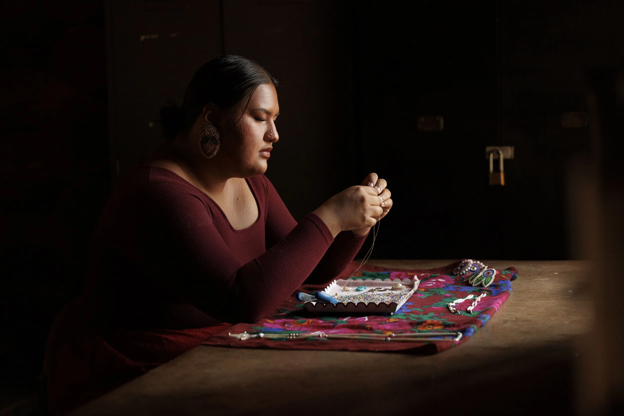 A woman with dark hair and earrings is sitting at a table, working on a craft project with beads and colorful fabric, in a dimly lit room.